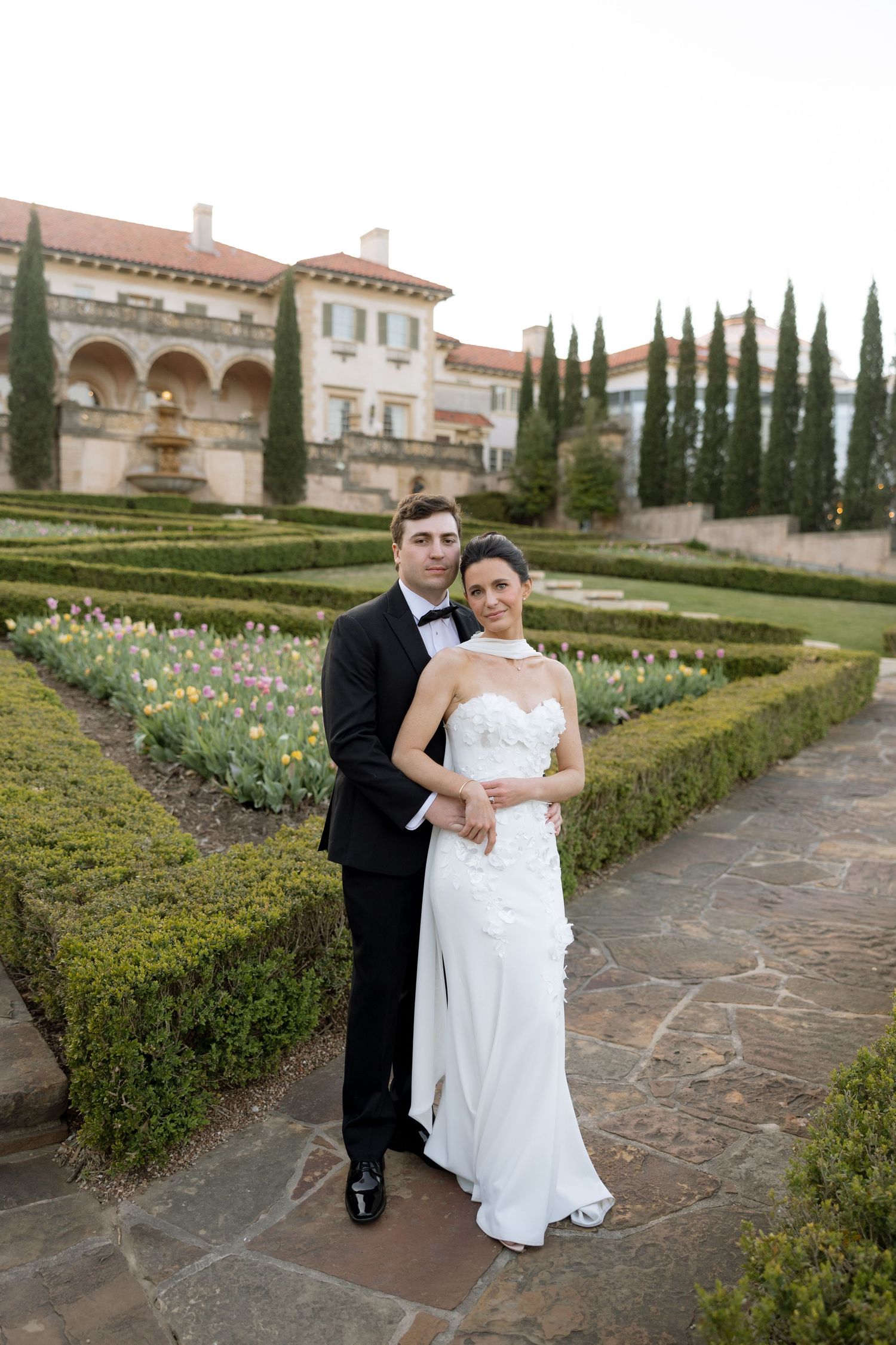 Bride and groom pose in formal attire in an elegant Italian garden with manicured hedges and a grand villa backdrop.