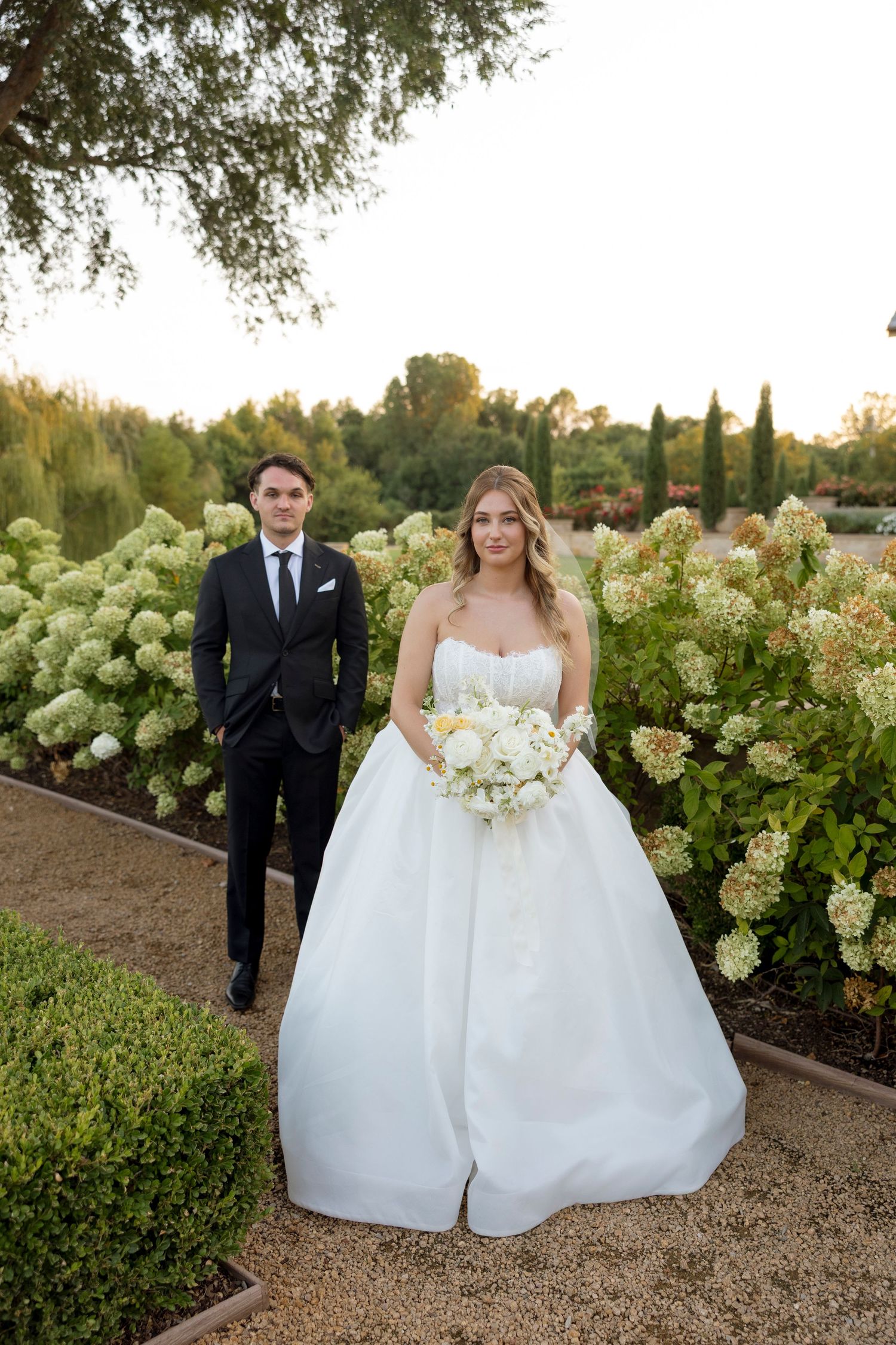 Bride in white ballgown holding bouquet with groom in black suit posing in garden with hydrangeas.