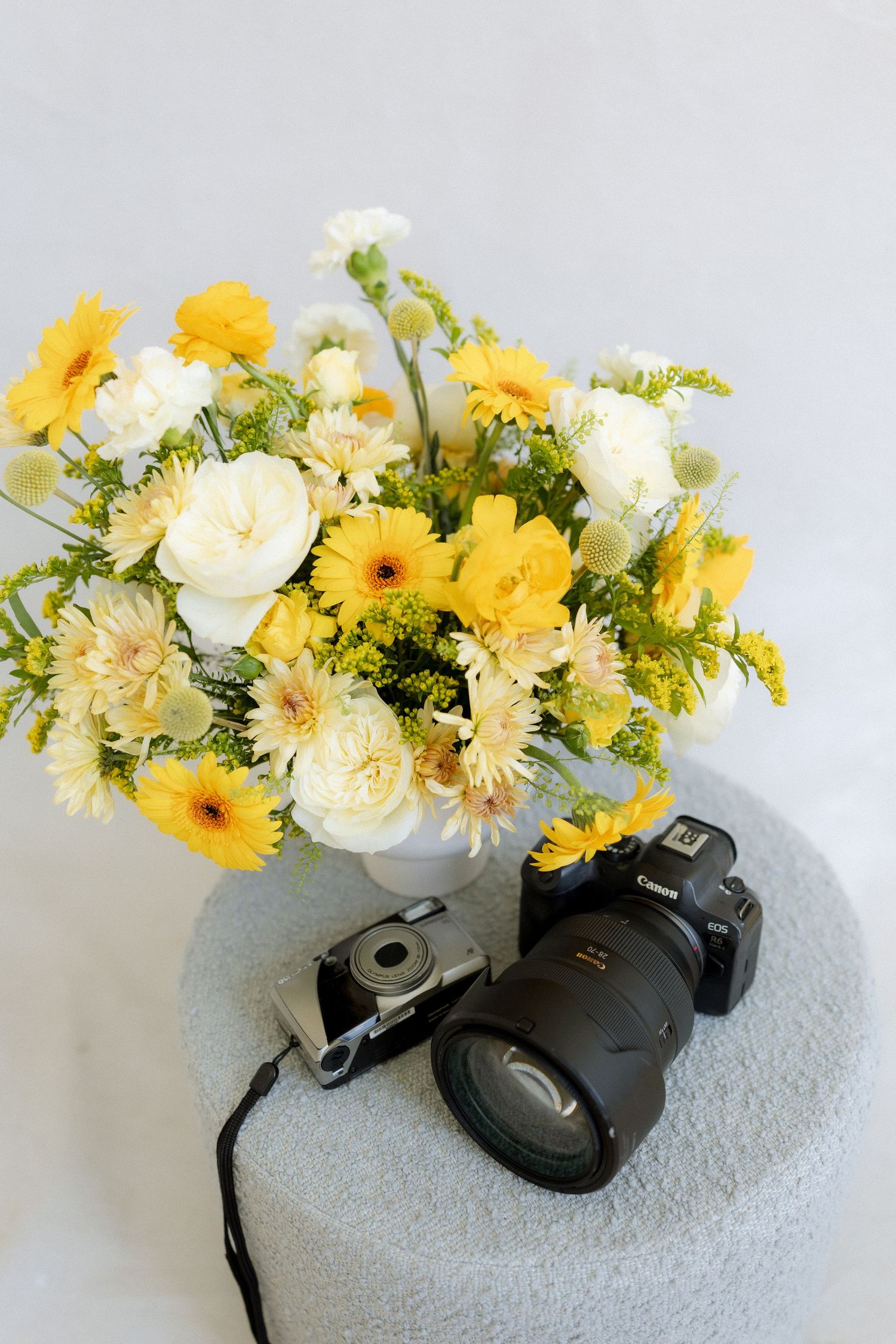 Yellow and white flower bouquet in vase beside Canon DSLR and film camera on a light blue ottoman.
