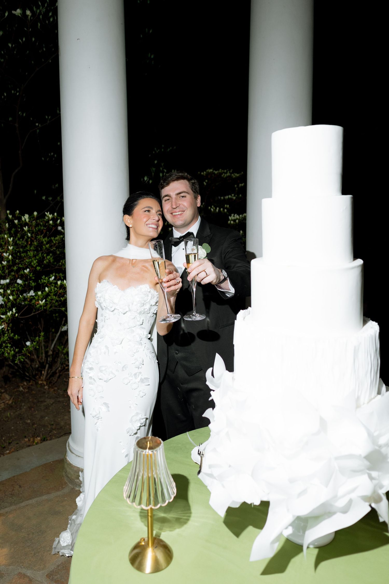 Bride and groom toasting champagne beside a tall white wedding cake outdoors at night near white columns.