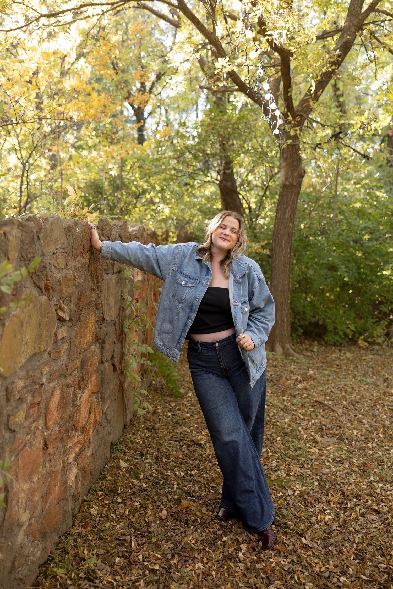 Person in denim jacket poses playfully against stone wall in autumn forest setting with golden leaves.