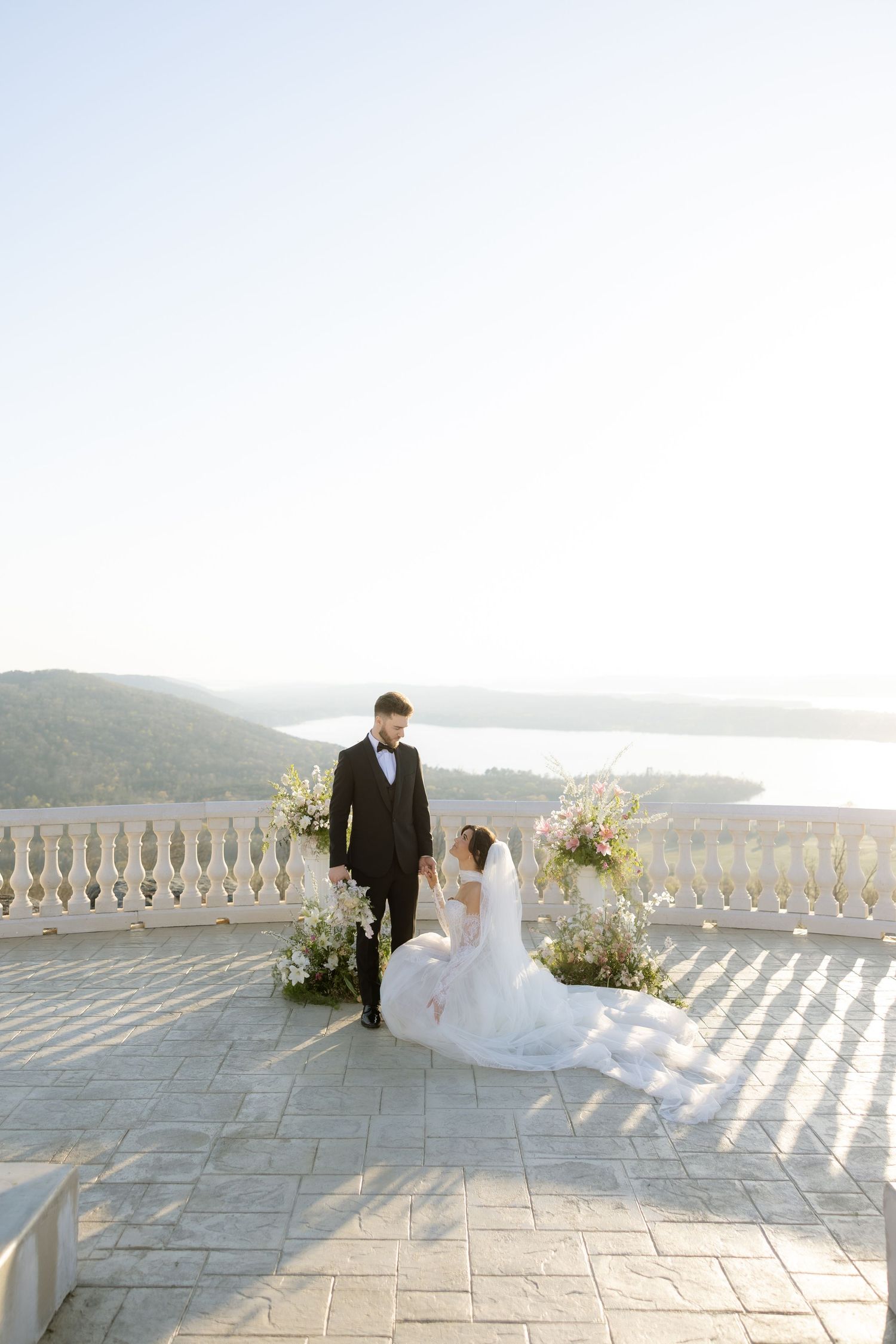 Bride in white gown and groom in black suit pose on elegant terrace with floral arch and scenic lake view.