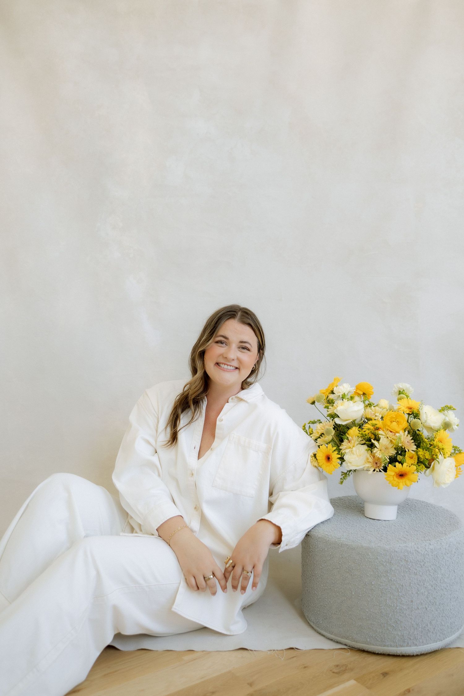Woman in white outfit sitting on floor beside yellow and white floral arrangement on gray ottoman.