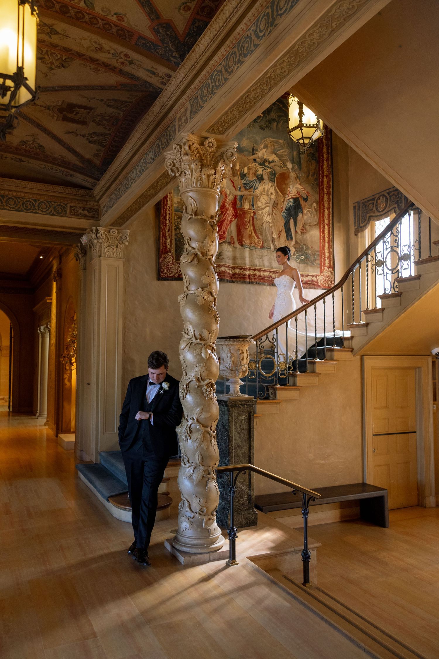 Groom in tuxedo checks phone beside ornate spiral staircase in grand historic venue with tapestry backdrop.