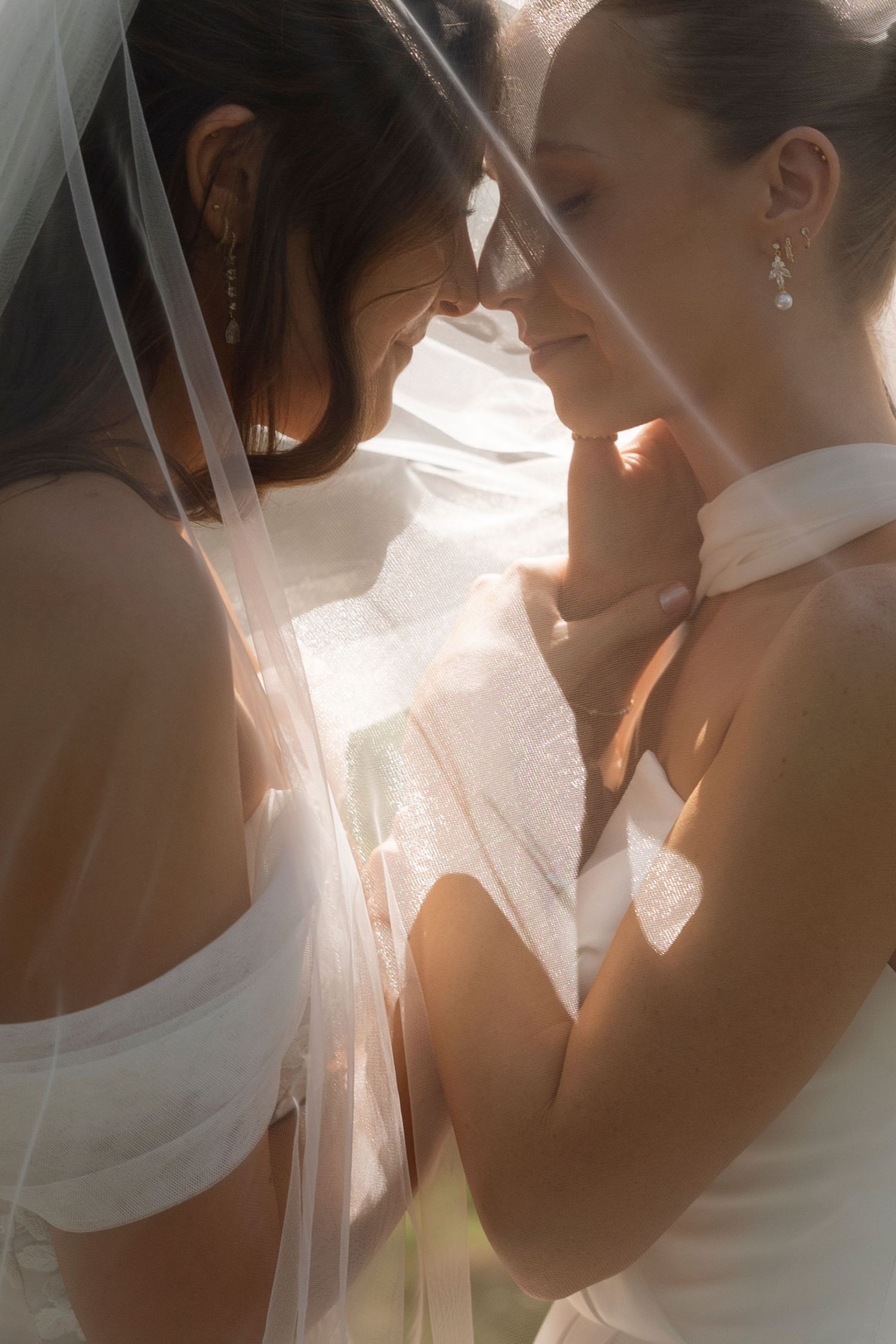 Two people in wedding attire share an intimate moment together in soft lighting with a delicate veil between them.