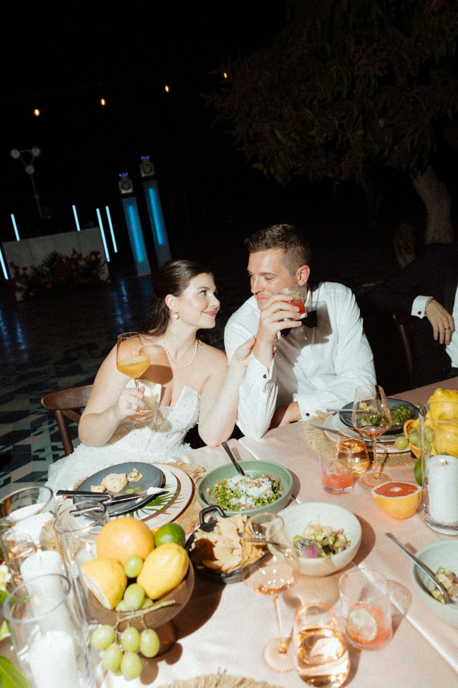 Bride and groom sharing a romantic toast at an outdoor wedding reception dinner table with elegant food and candles at night.