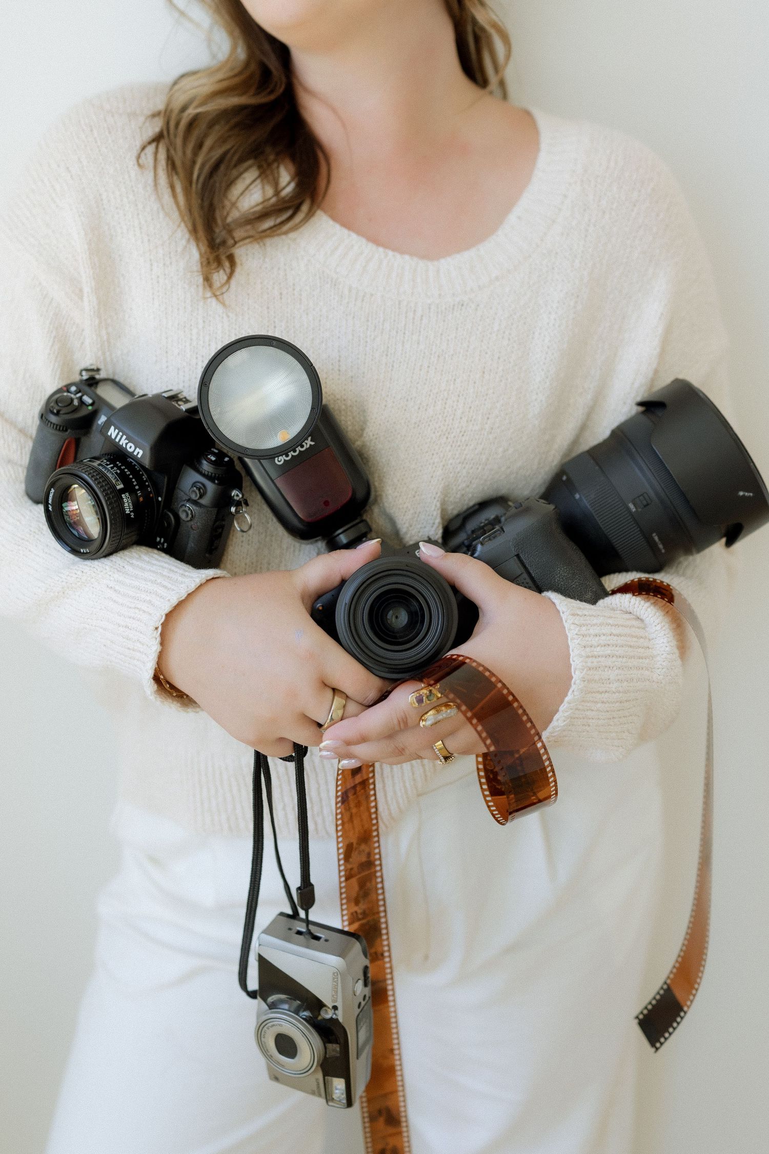 Woman in white sweater holding multiple cameras including a Nikon DSLR, flash, lenses, and vintage film camera with brown strap.