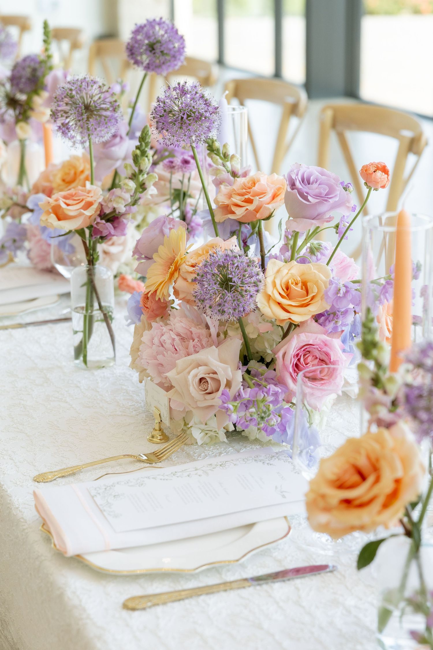 Elegant wedding table with pastel floral centerpieces featuring peach roses, purple allium, and pink blooms with gold cutlery.