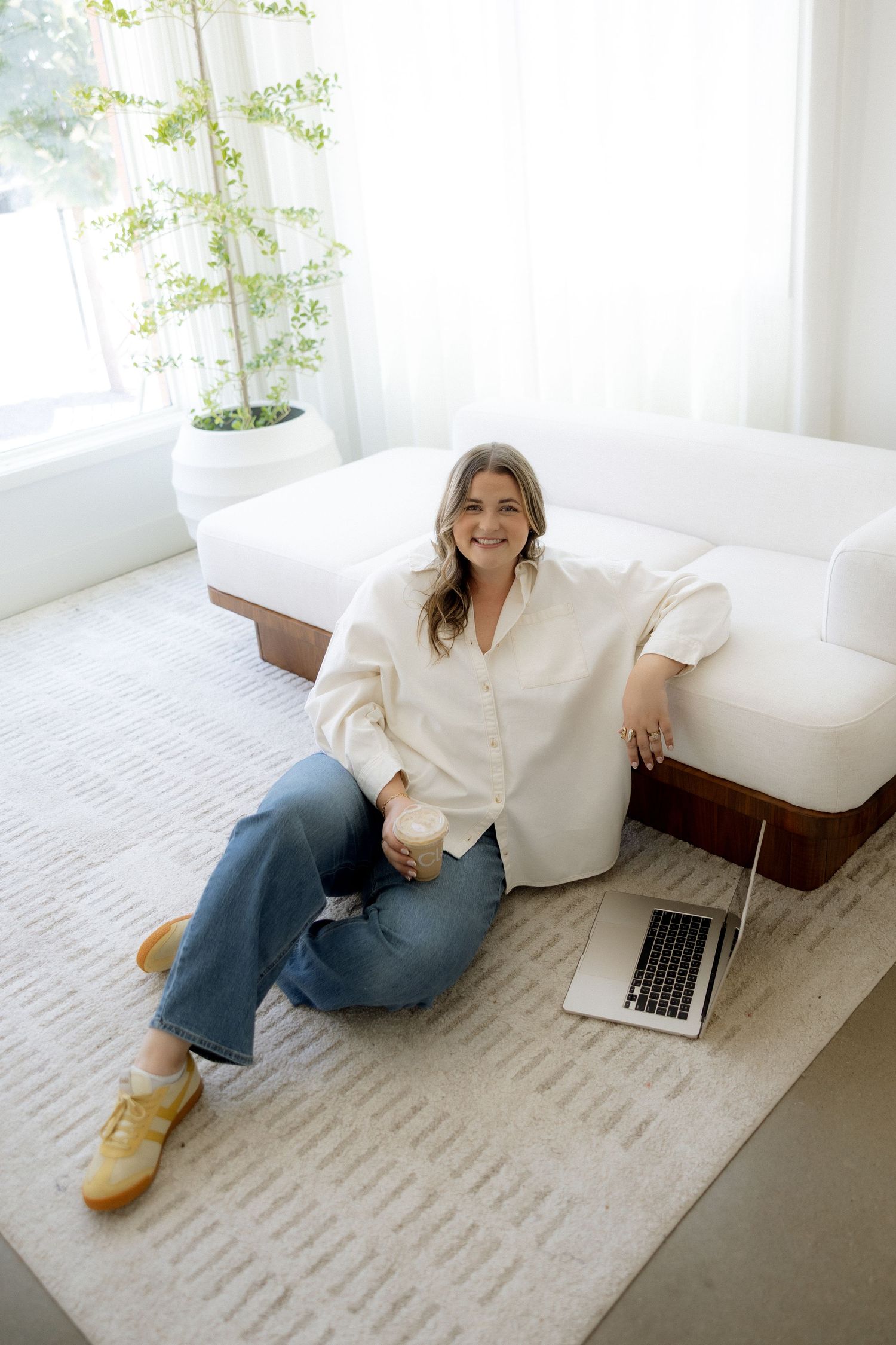 Woman in white shirt and jeans sits on floor leaning against white sofa with laptop in bright modern living room.