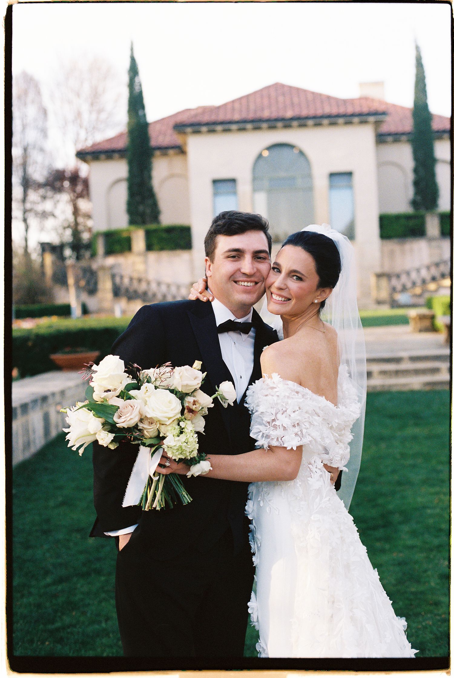 Bride in off-shoulder floral gown holding white bouquet poses with groom in tuxedo before elegant Italian-style villa.