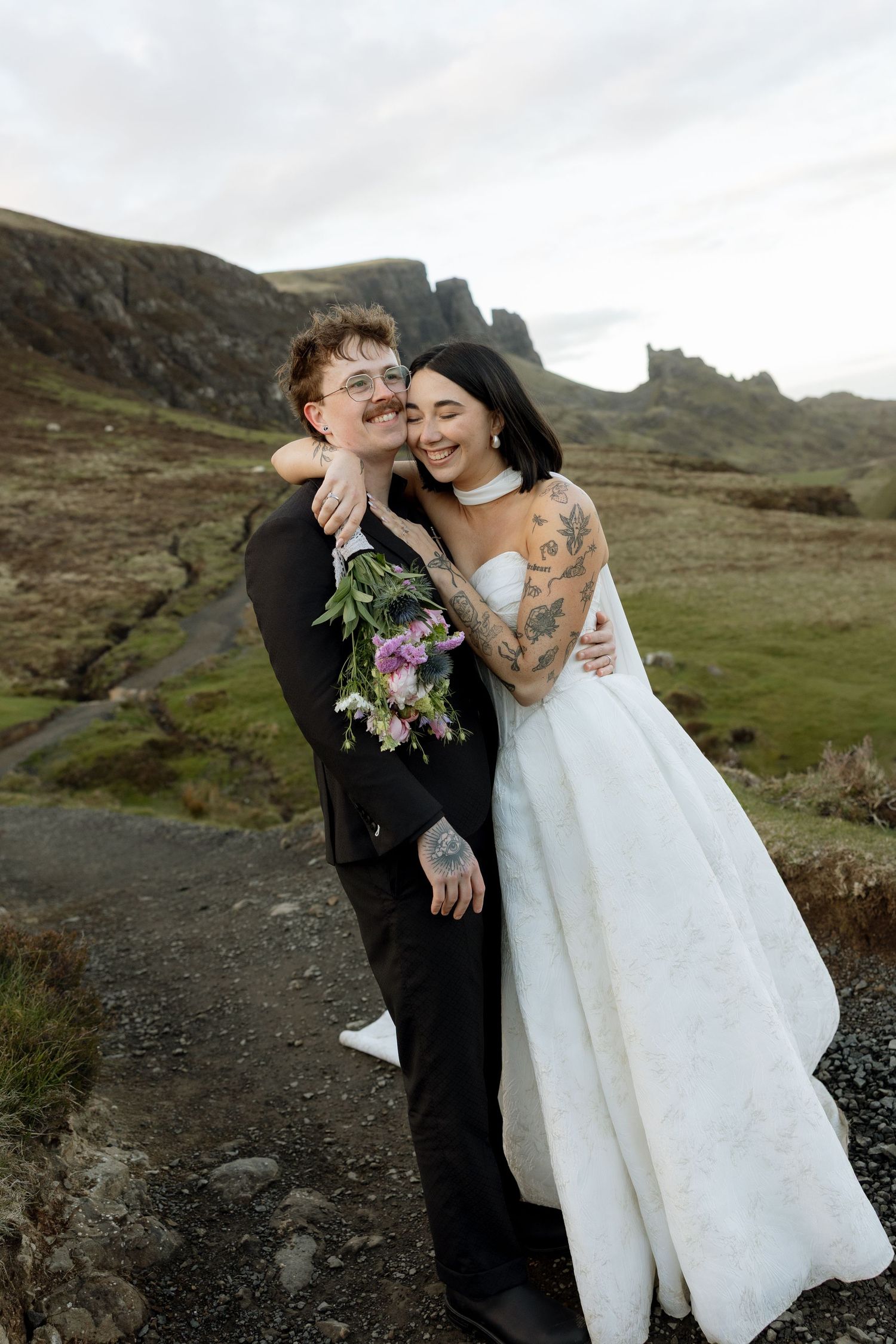 Tattooed couple in wedding attire embrace outdoors in Scottish Highlands, bride in white gown holding bouquet.