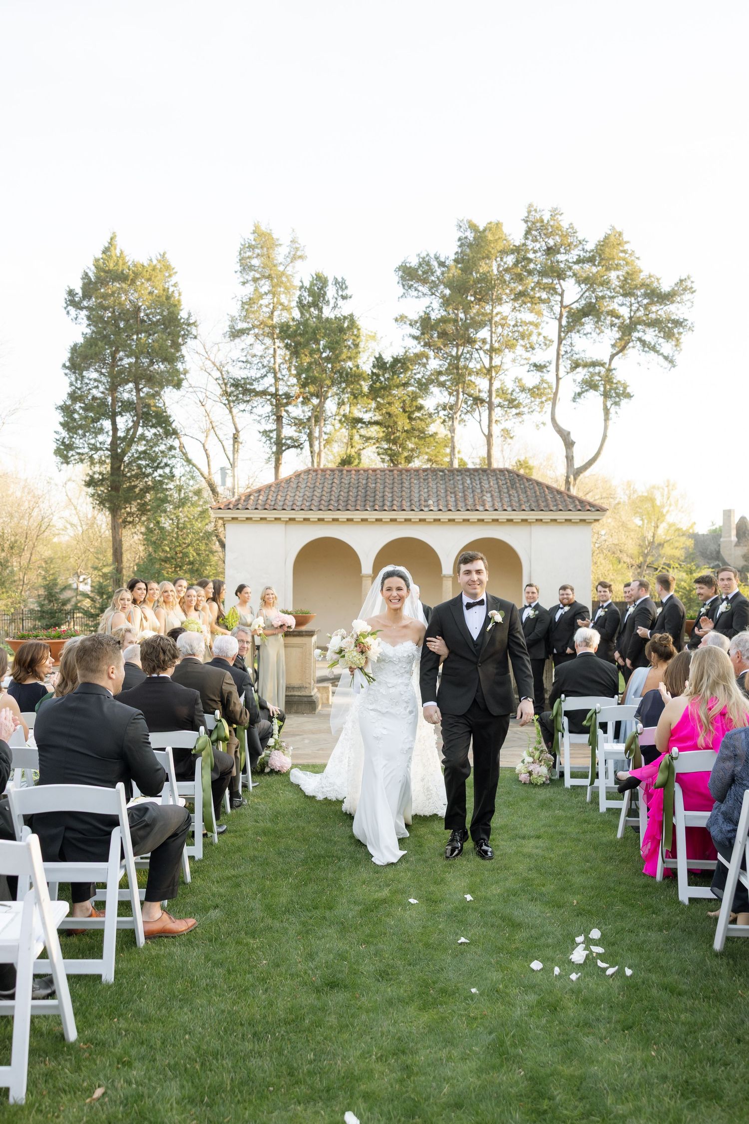 Bride and groom walk down outdoor aisle after wedding ceremony, guests seated in white chairs, gazebo backdrop.