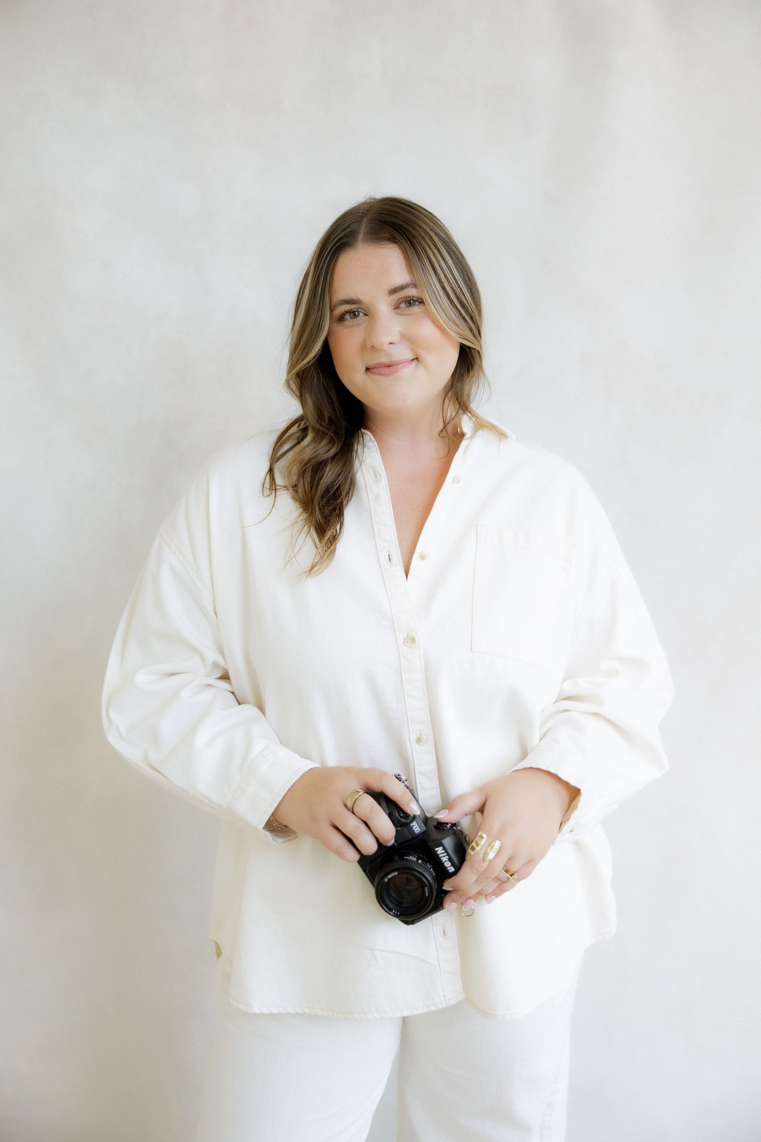 A smiling woman in a white oversized shirt holds a camera against a light gray background.
