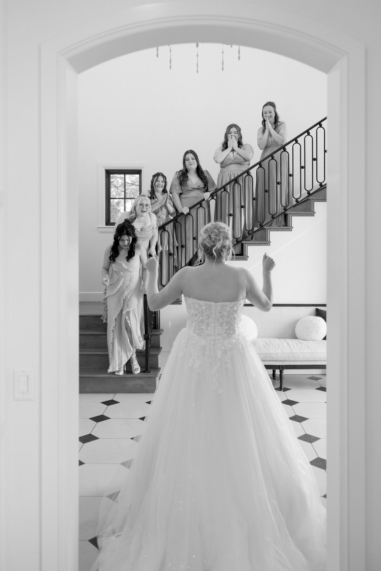Bride in strapless ball gown reveals herself to bridesmaids on staircase in black and white wedding photo.