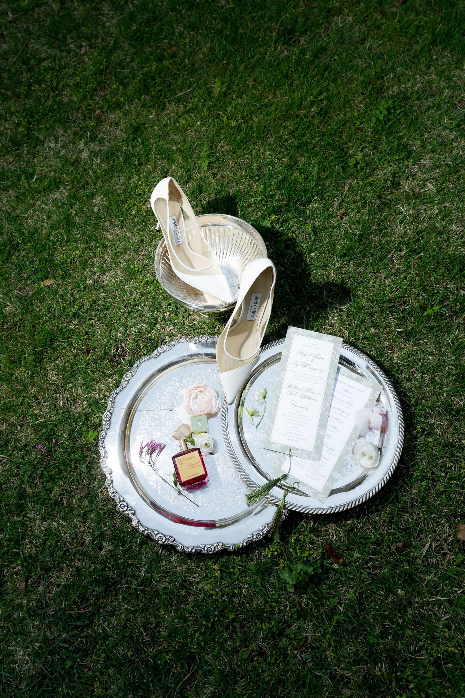 Silver trays on grass displaying bridal accessories including heels, ring box, flowers, and wedding stationery.