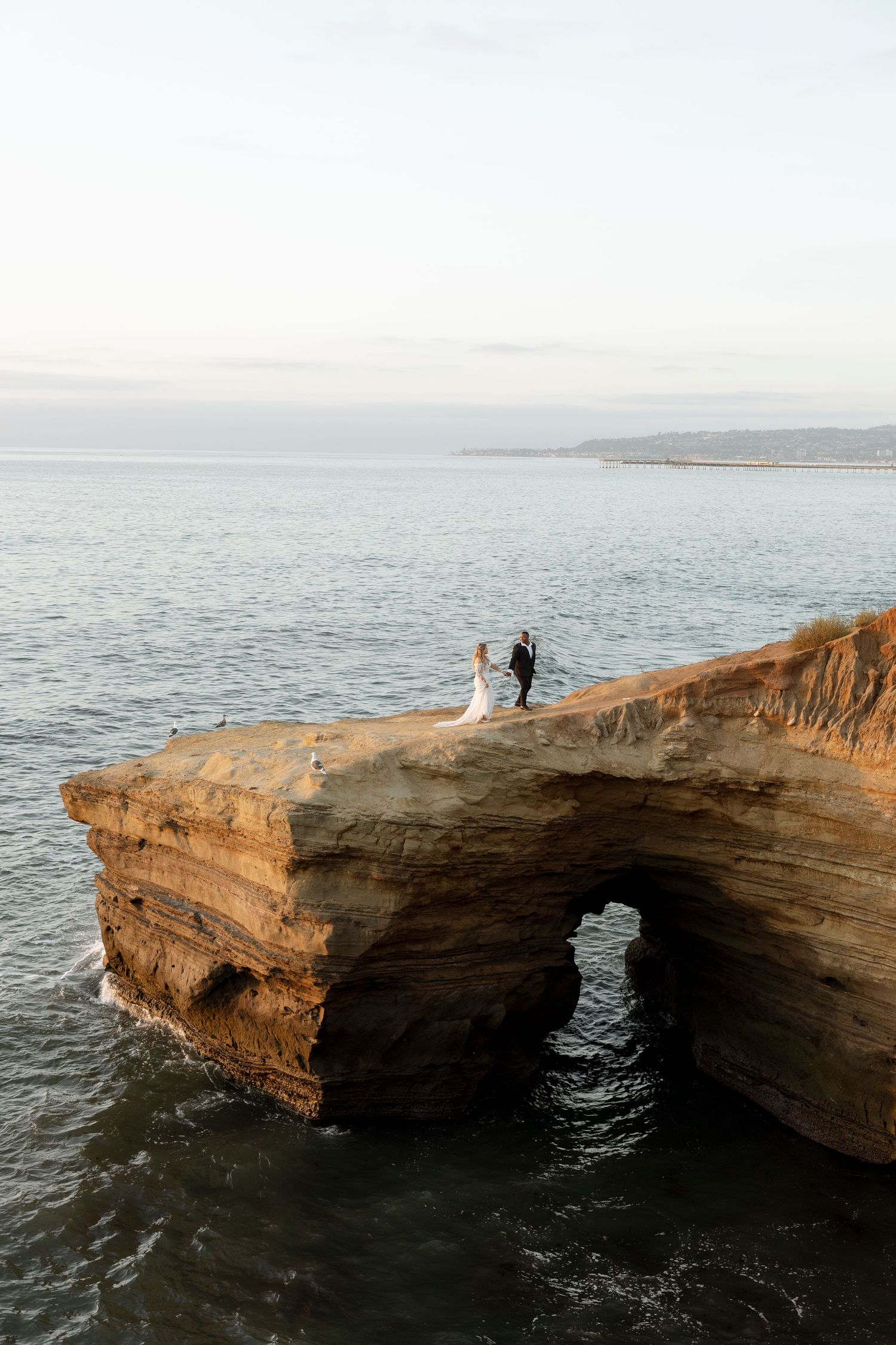 Two silhouettes stand atop a dramatic natural stone arch formation over the ocean at sunset in San Diego.