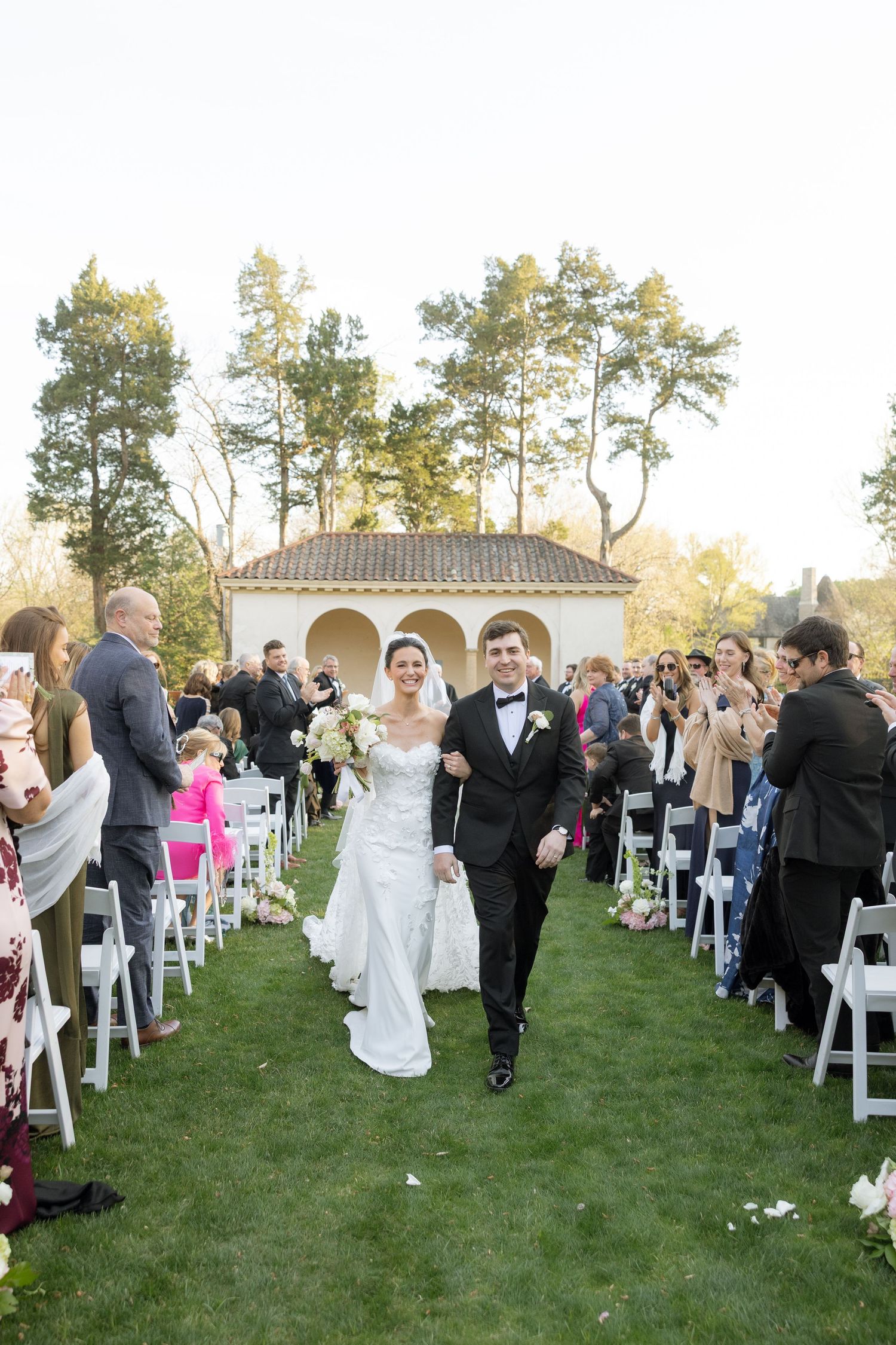 Bride and groom walk down outdoor wedding aisle lined with white chairs and guests, with a garden pavilion backdrop.
