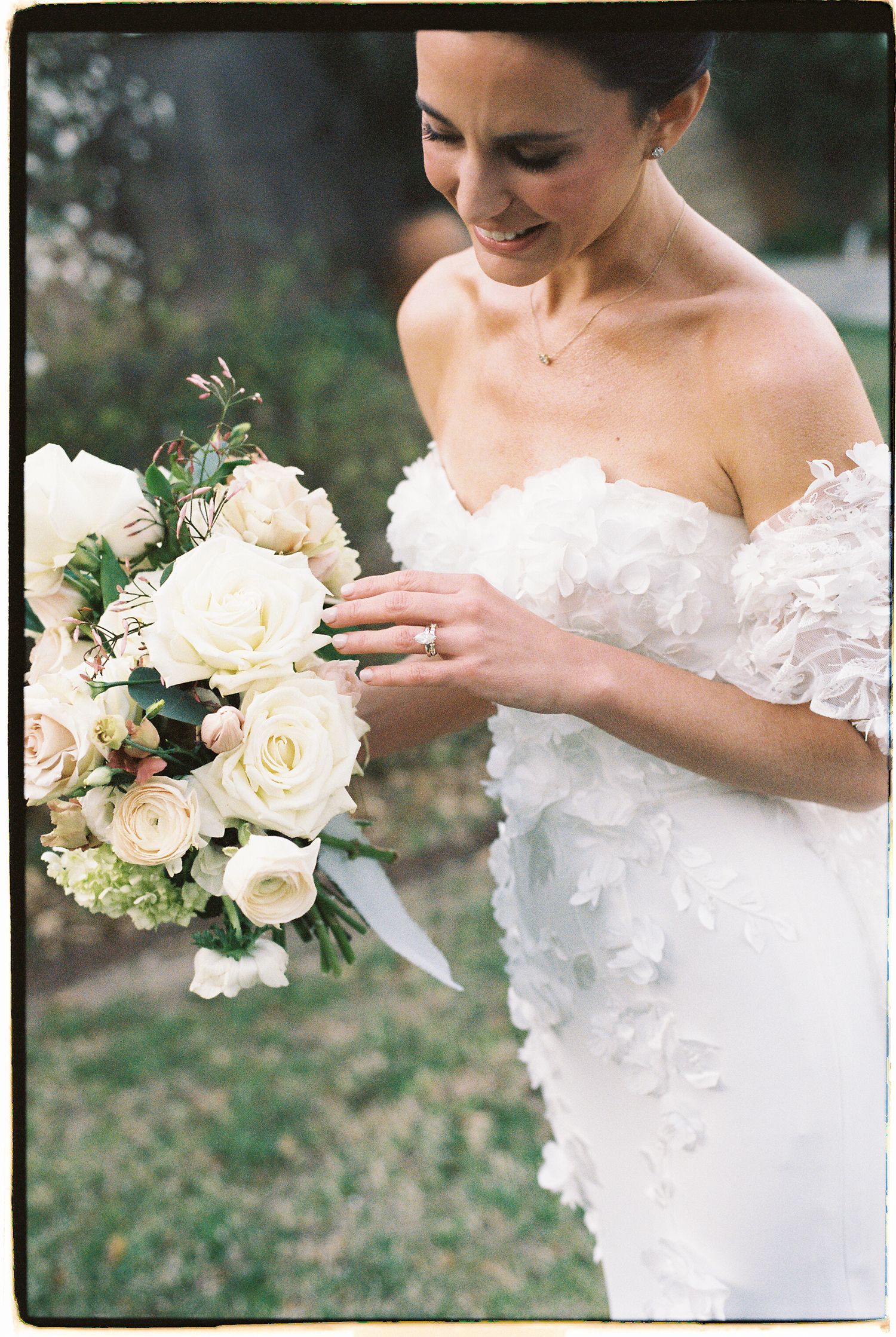 Smiling bride in a floral off-shoulder gown holds a lush white rose and ranunculus bouquet outdoors.