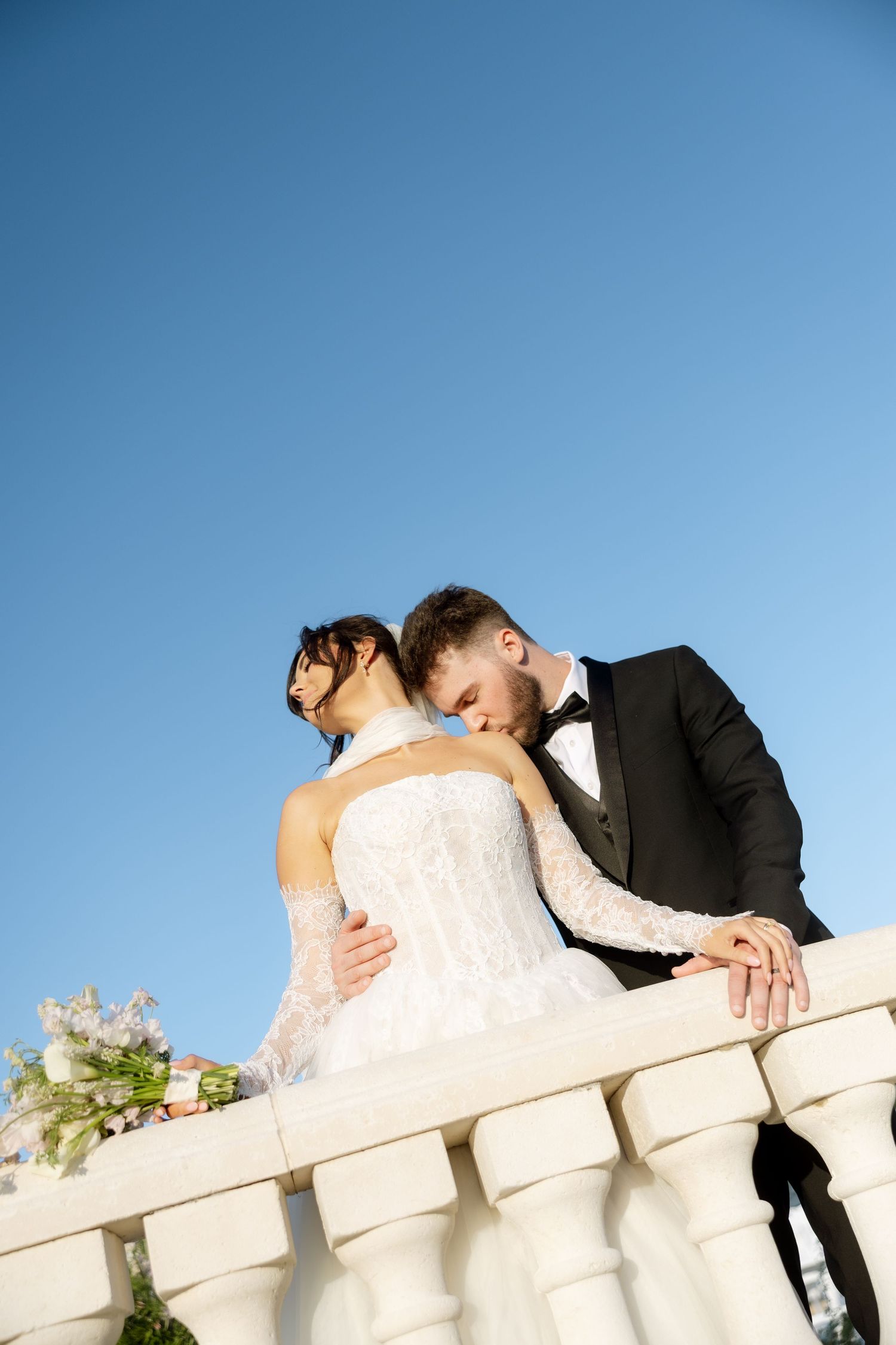 A groom in a black tuxedo tenderly kisses his bride in a white lace gown on a sunlit balcony under a clear blue sky.