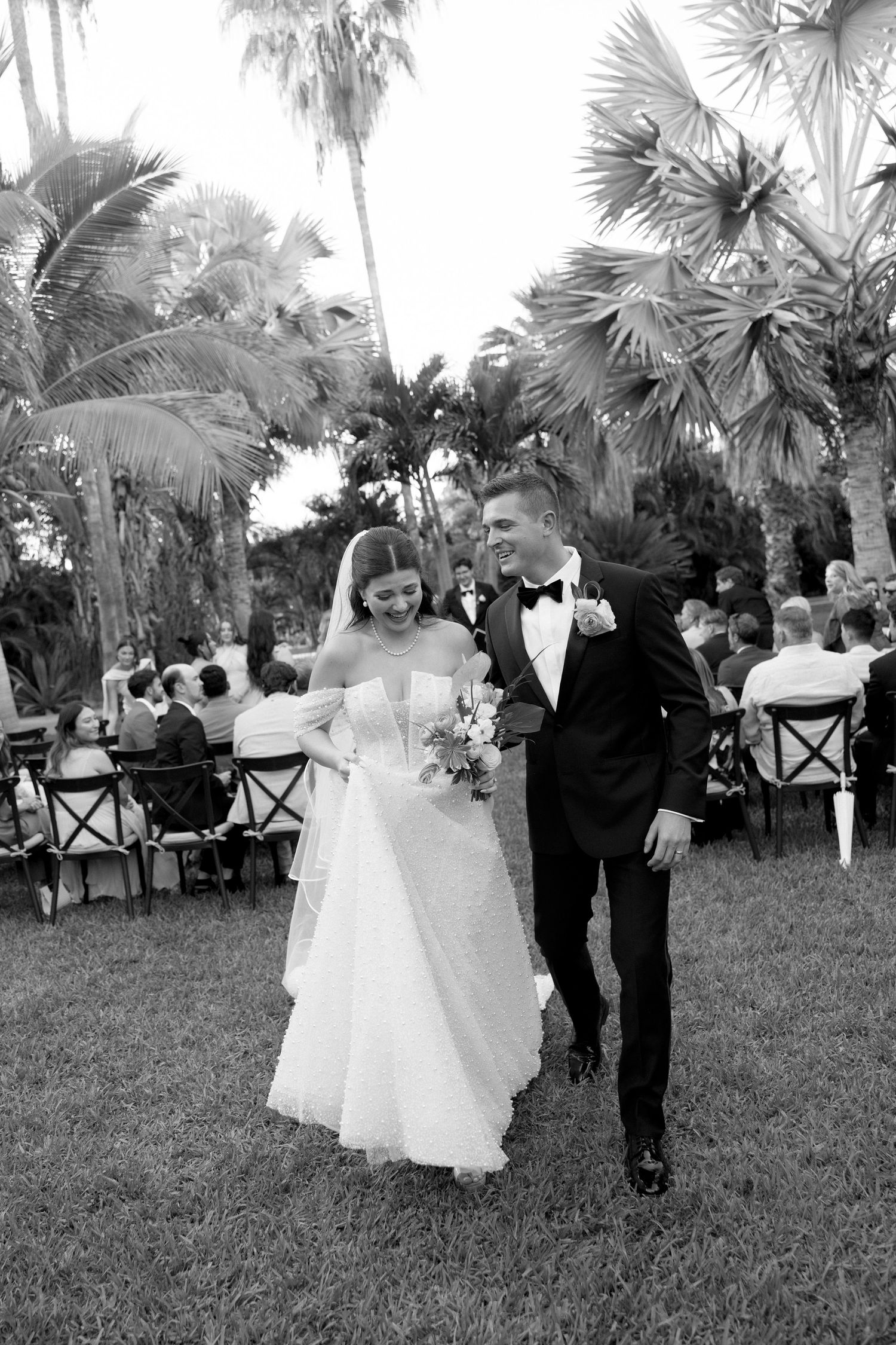 Black and white photo of a smiling bride and groom walking down the aisle at an outdoor wedding ceremony surrounded by palm trees.