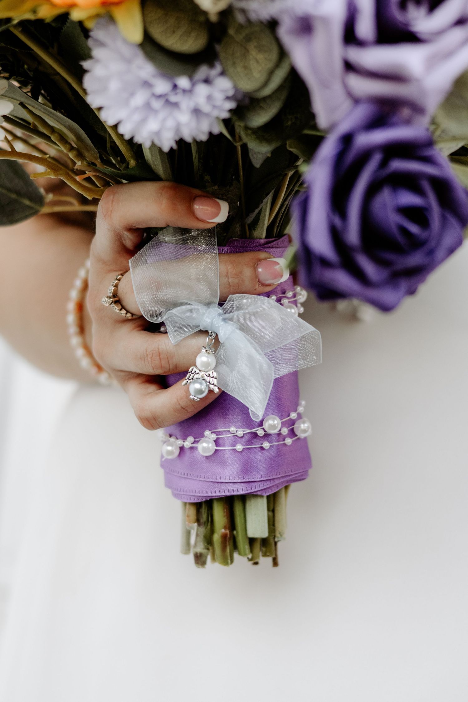 Bride's hand holds a purple rose bouquet with a lavender ribbon stem wrap, pearl strands and an angel charm against a white dress.