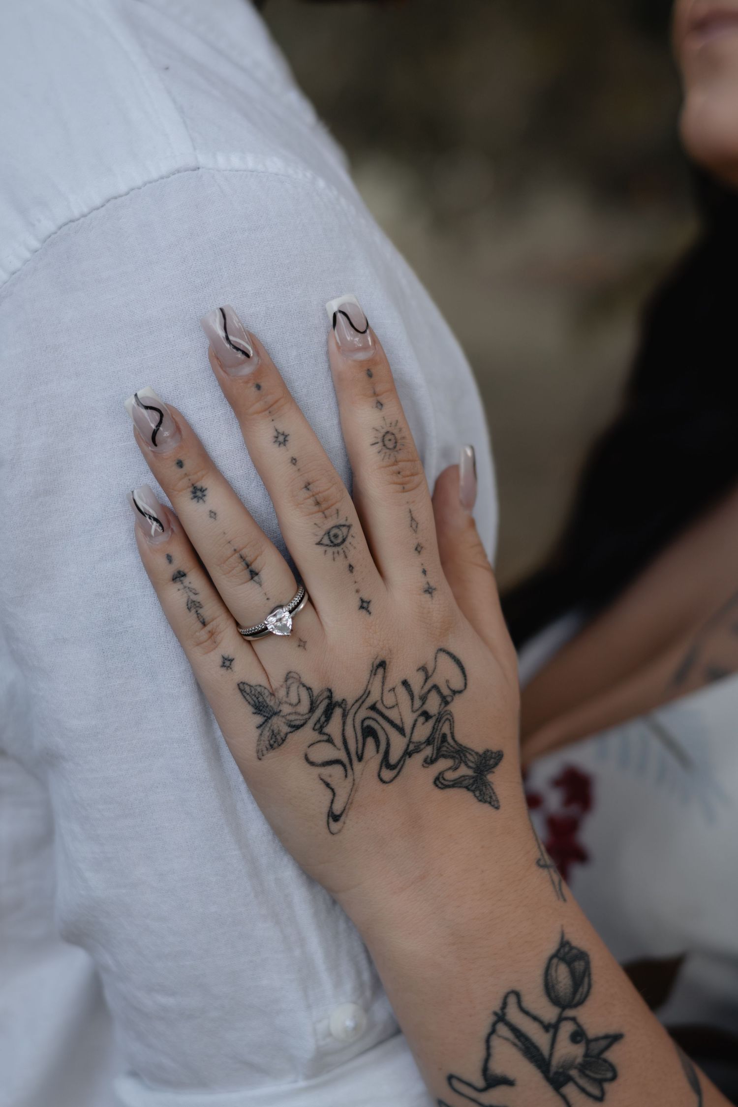 Tattooed hand with a diamond engagement ring and wavy black-tipped nails resting on a person's white linen shirt.