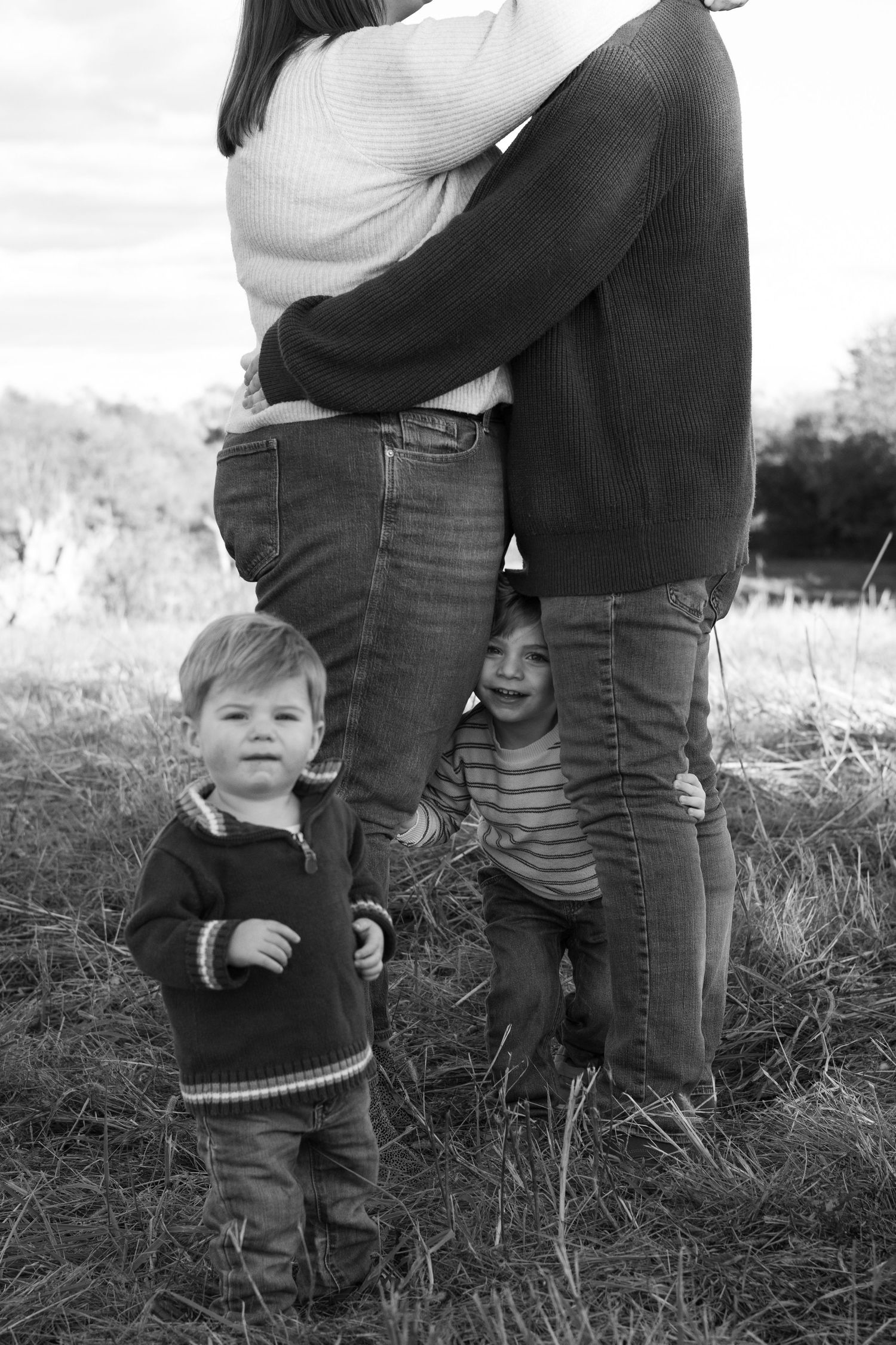 Black and white family photo of two parents embracing in a grassy field with two young children standing close beside their legs.