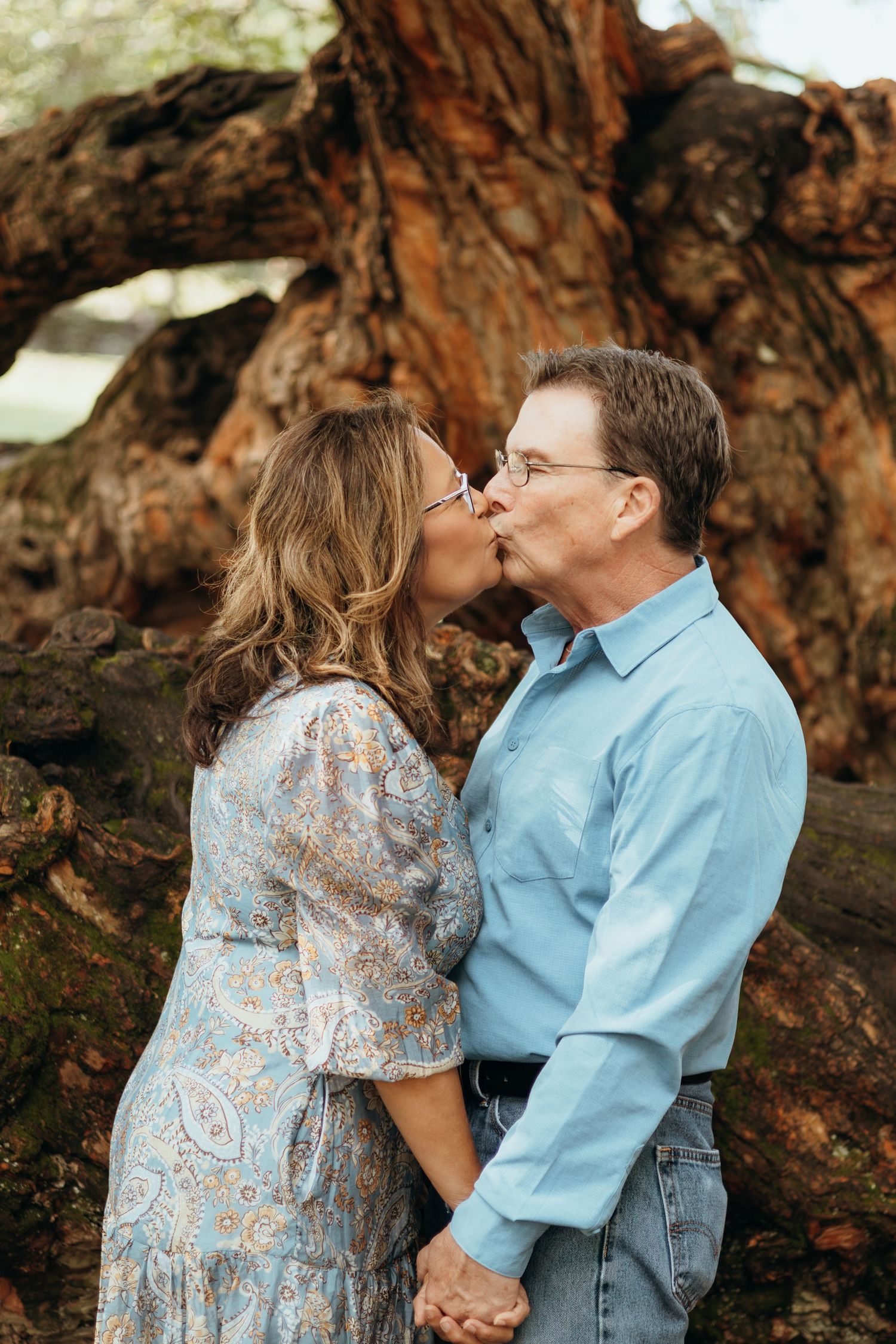 Couple sharing a kiss and holding hands in front of a large gnarled oak tree, she in a floral dress and he in a blue button-up shirt.