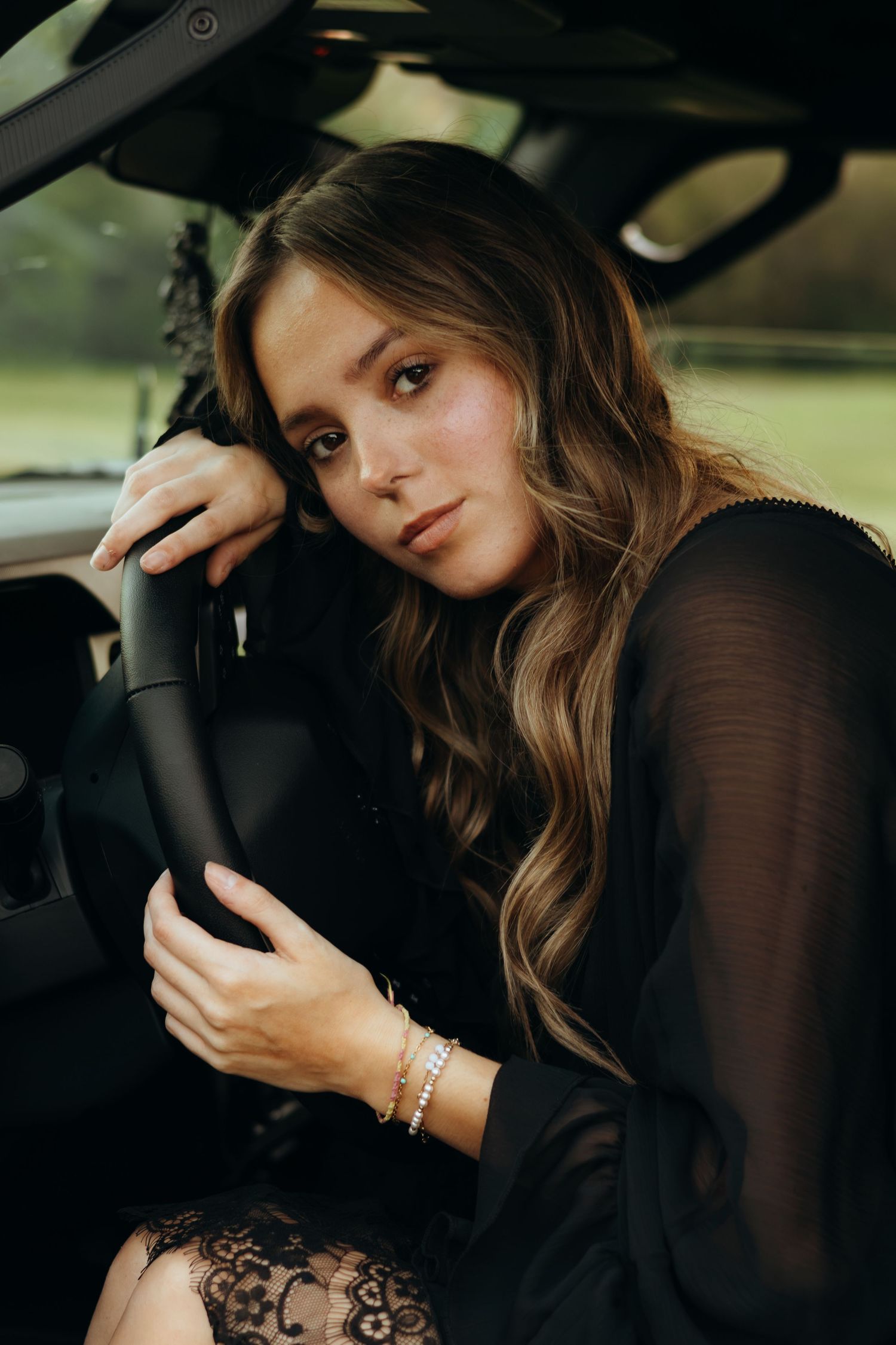 Young woman with long wavy hair in a black lace outfit leans her head on her hand while sitting in a vehicle.