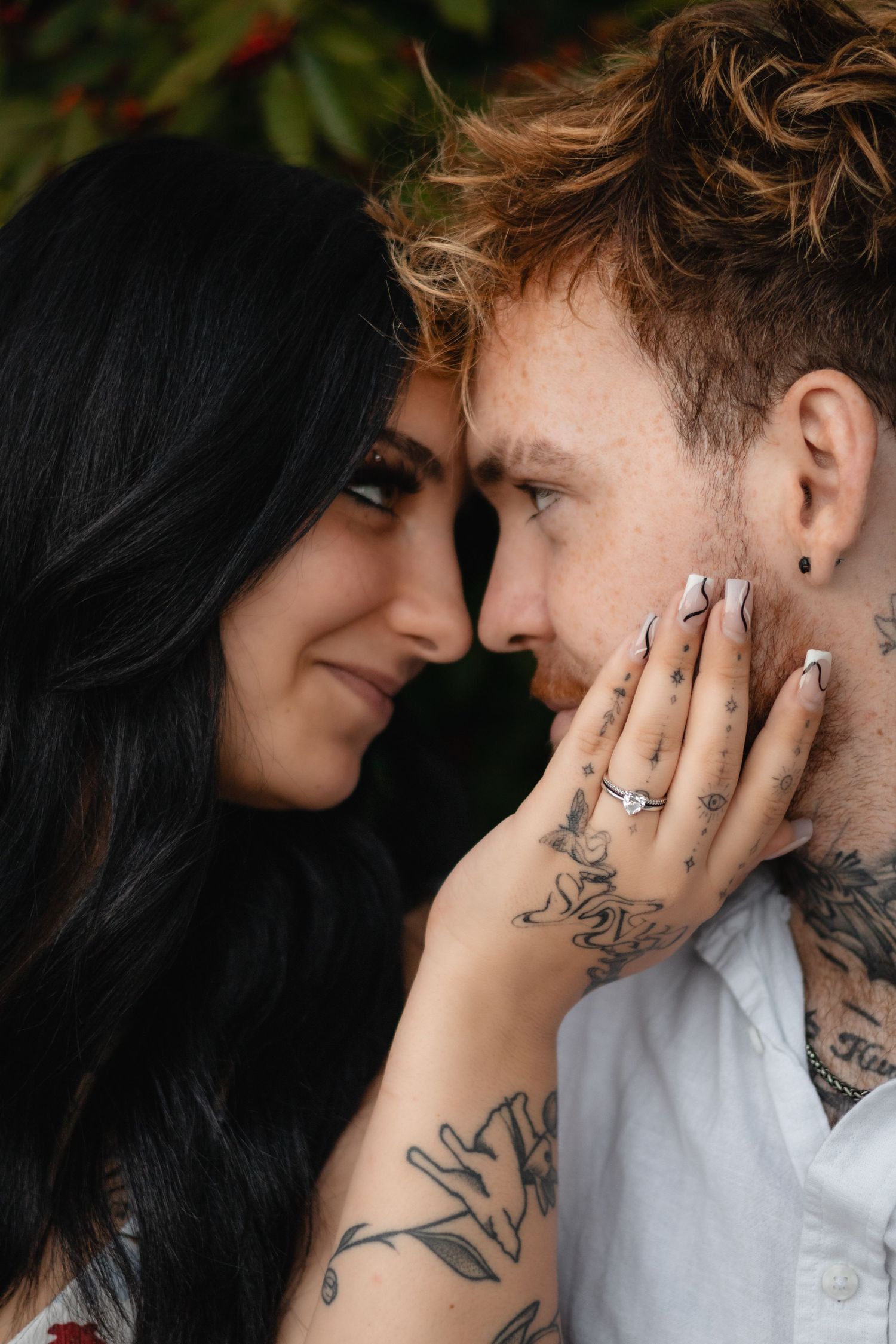 Tattooed couple gazing at each other closely as the woman with long dark hair touches his cheek, showing her engagement ring.