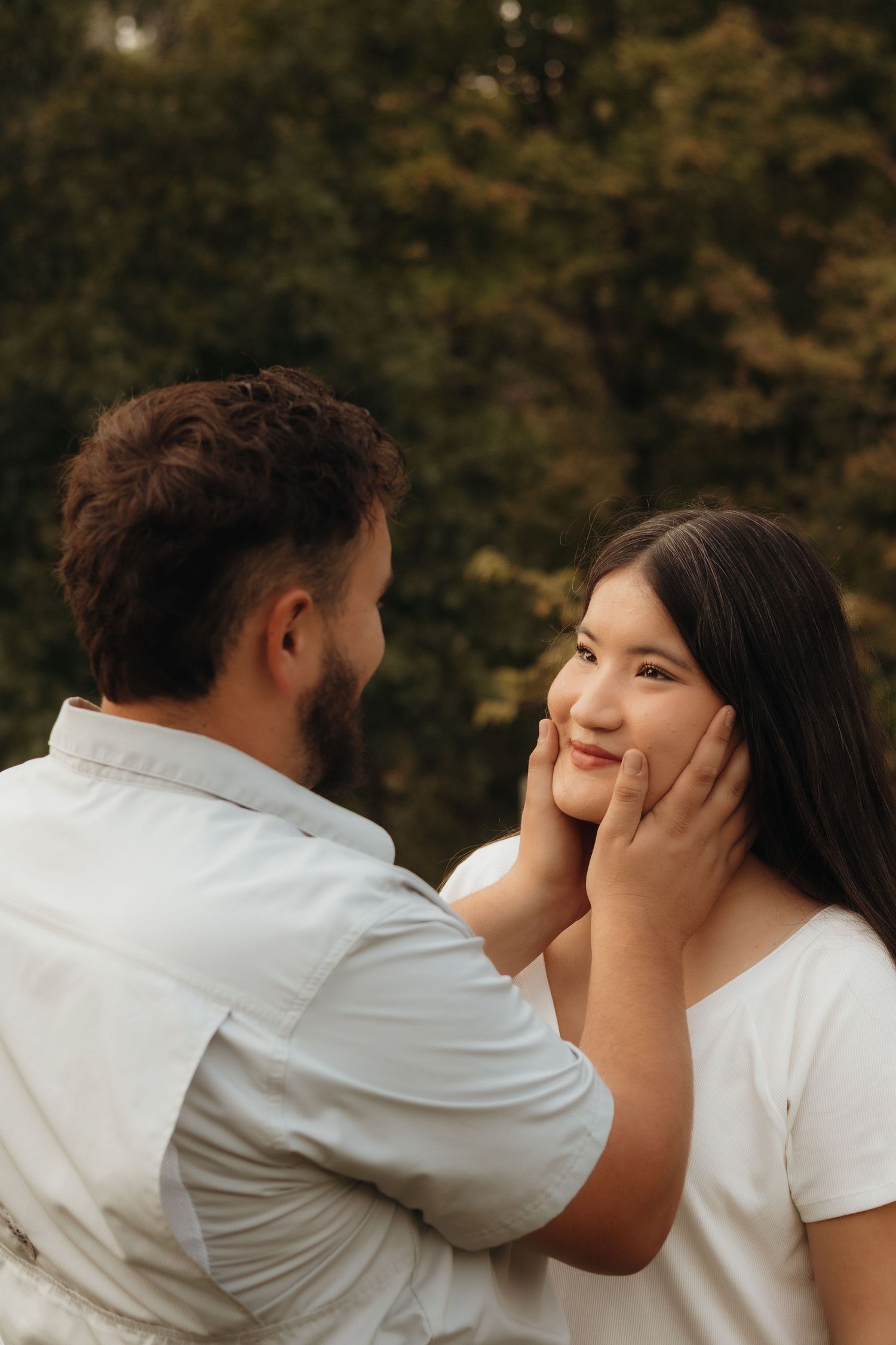 Smiling woman with long dark hair gazes up as a bearded man in a light button-up shirt gently cups her face outdoors among green trees.