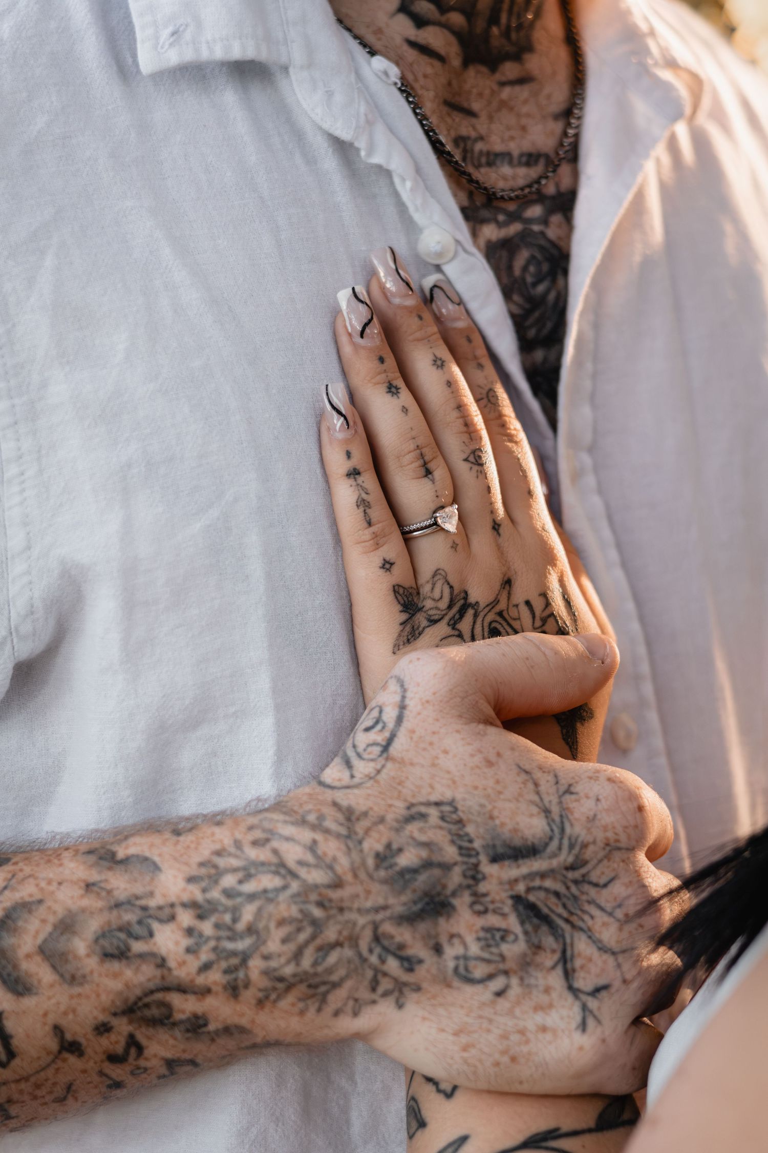 Close-up of a tattooed couple embracing, her hand with a diamond engagement ring and manicured nails resting on his white shirt.
