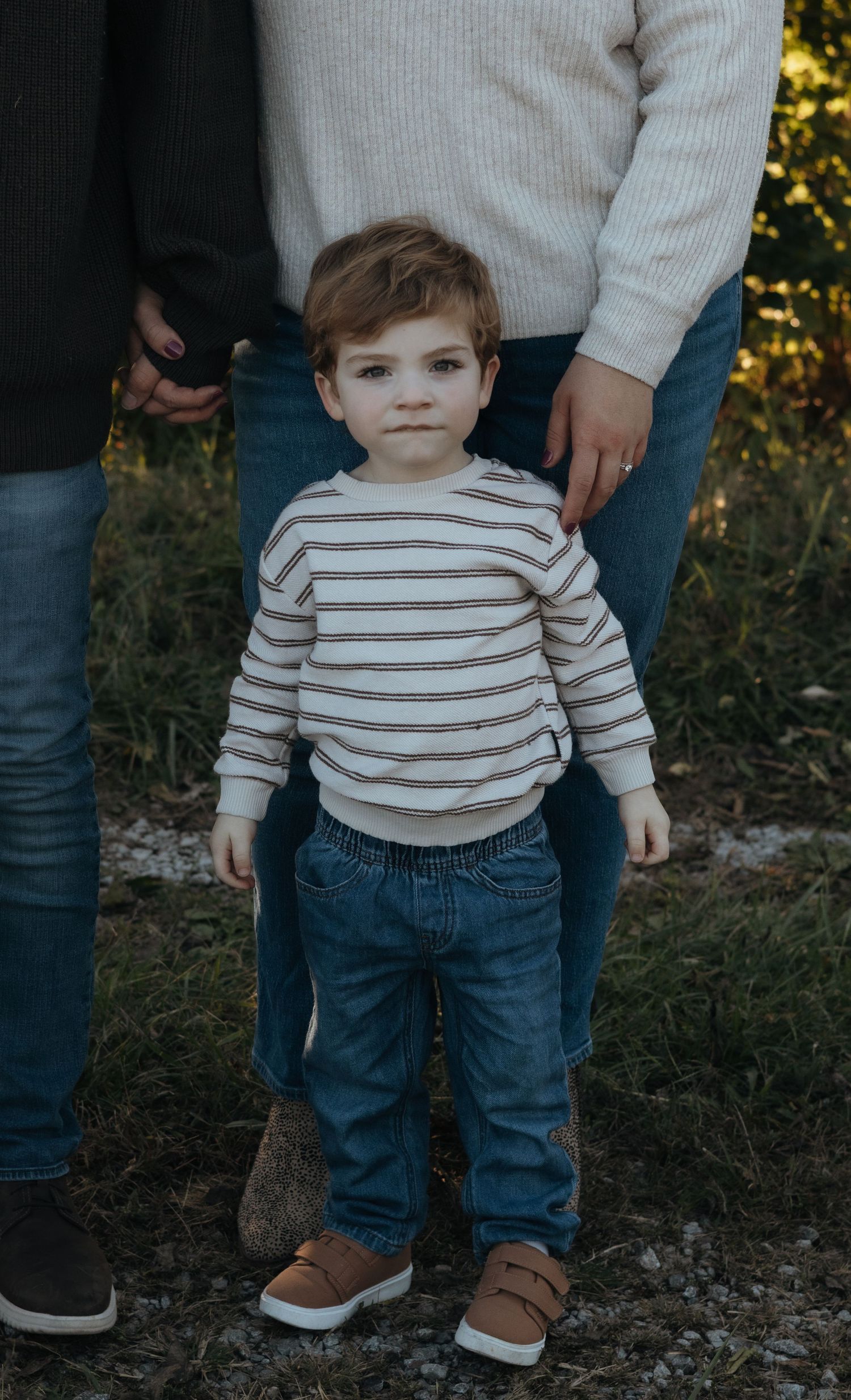 Toddler boy in a striped sweater, jeans and brown sneakers stands on grass between two adults in an outdoor family portrait.