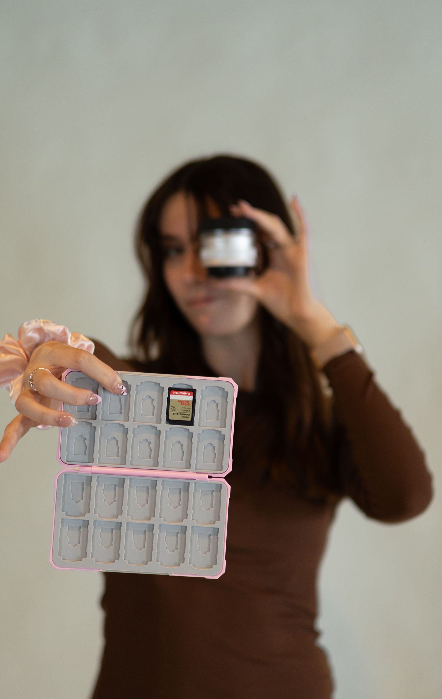 Woman in a brown top holds up a pink game card case toward the camera while looking through a lens, blurred behind.