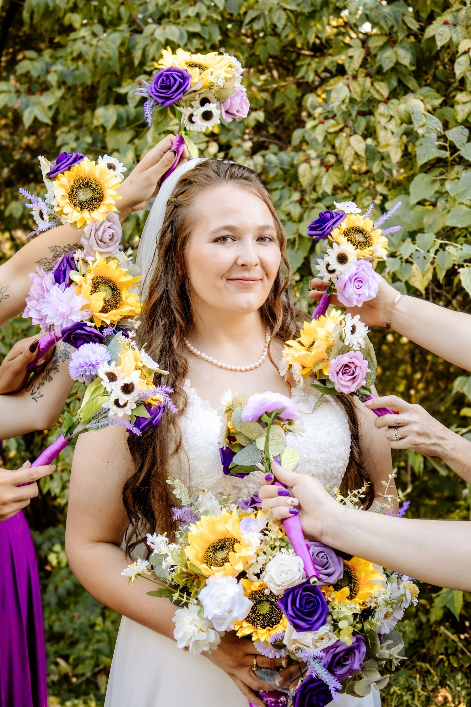 Smiling bride in a lace gown and pearl necklace surrounded by bridesmaids' hands holding sunflower and purple floral bouquets outdoors.