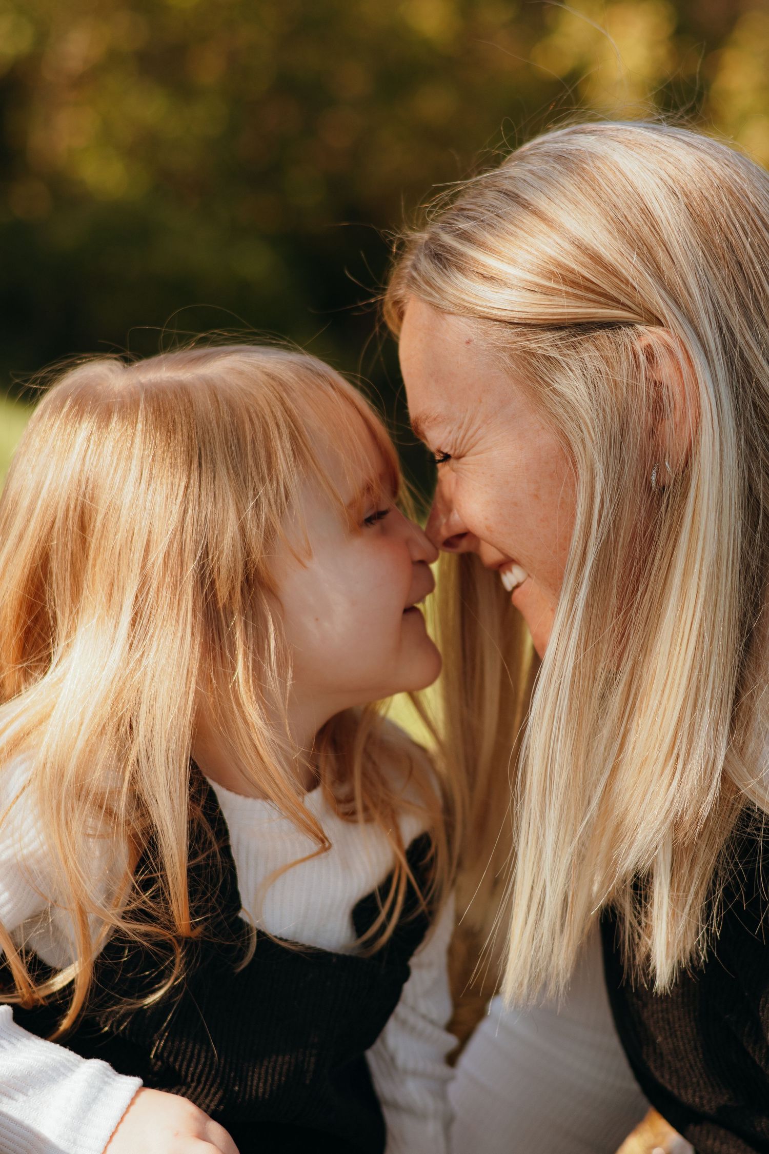 Smiling woman and young girl with long blonde hair touch noses in a tender moment outdoors, surrounded by warm, softly blurred autumn light.