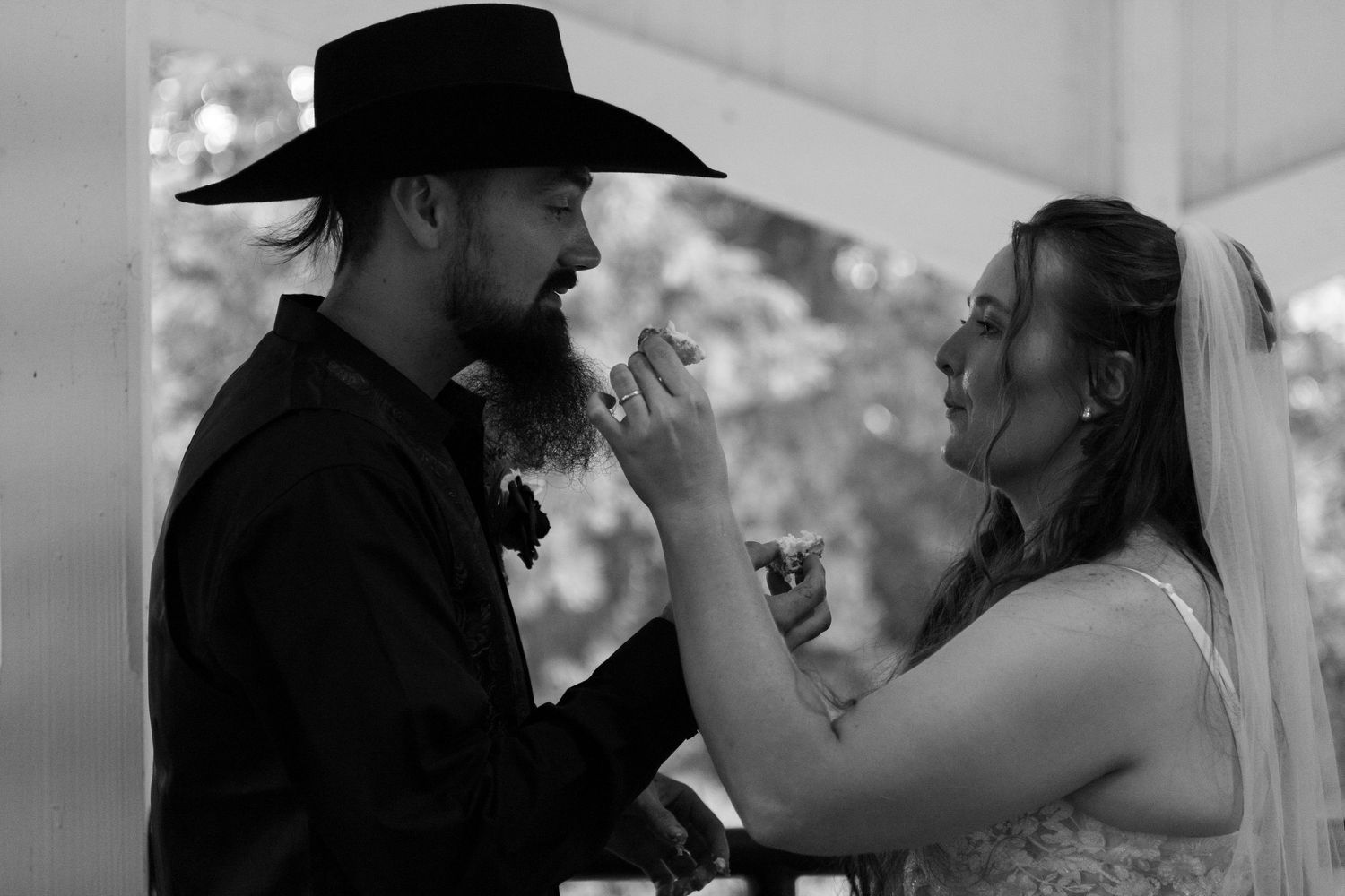 Black and white photo of a bride in a veil feeding wedding cake to a bearded groom in a cowboy hat during a rustic outdoor ceremony.