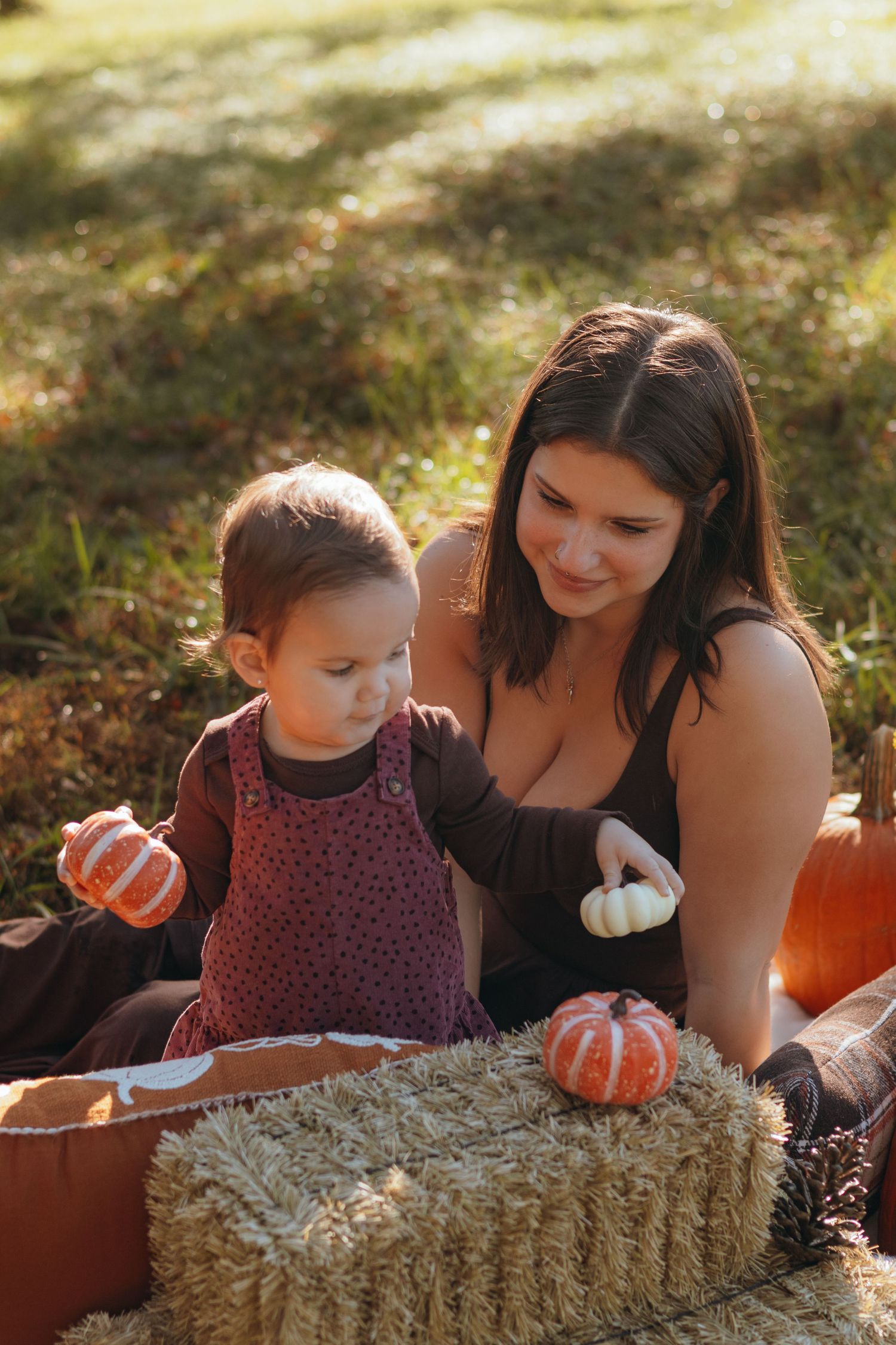 A smiling woman and a toddler in a burgundy pinafore sit on a hay bale holding small pumpkins in a sunny autumn field.