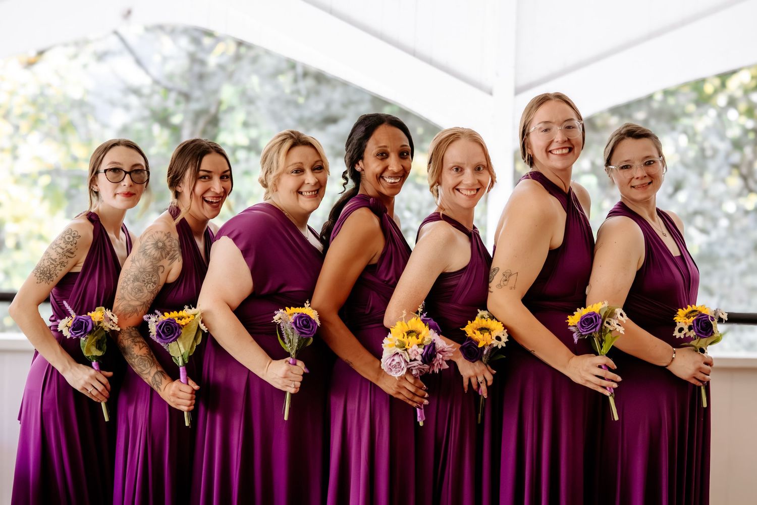 Seven smiling bridesmaids in matching plum purple dresses stand in a row holding small sunflower and purple wildflower bouquets.