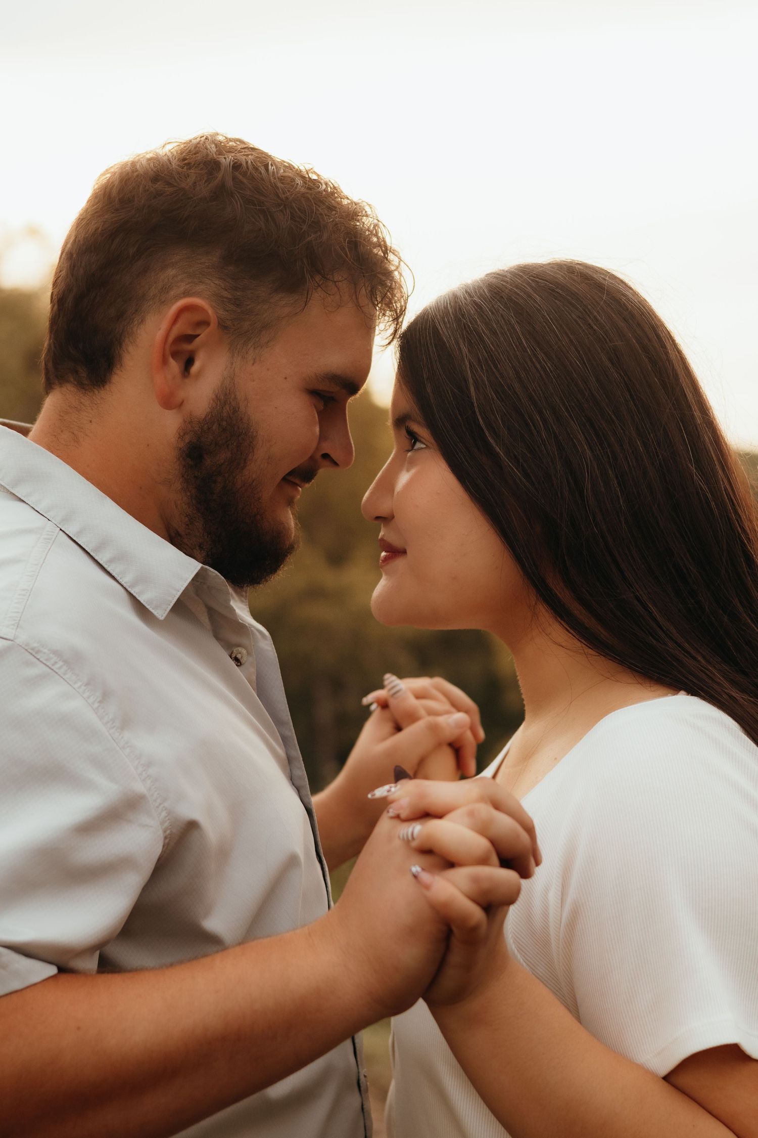 Romantic couple holding hands and gazing into each other's eyes outdoors during a soft golden hour engagement photoshoot.