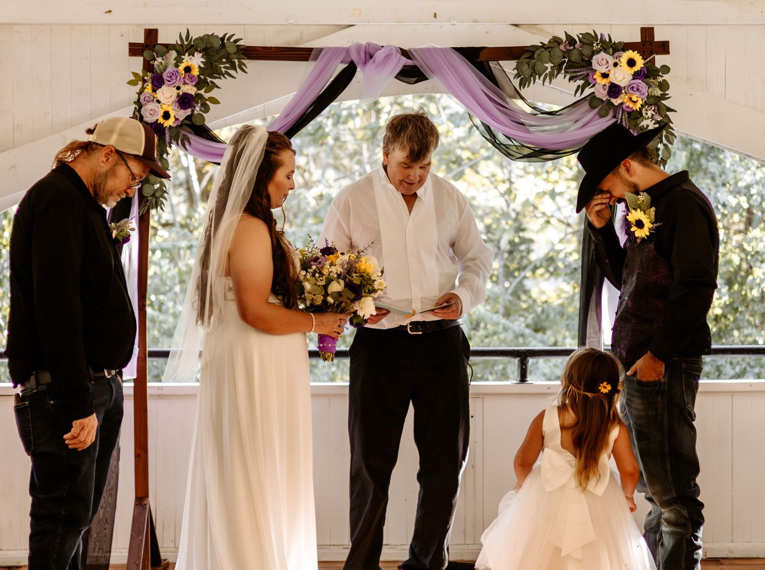 Bride in white gown holding a bouquet stands before an officiant under a floral wedding arch with a flower girl and groomsmen nearby.