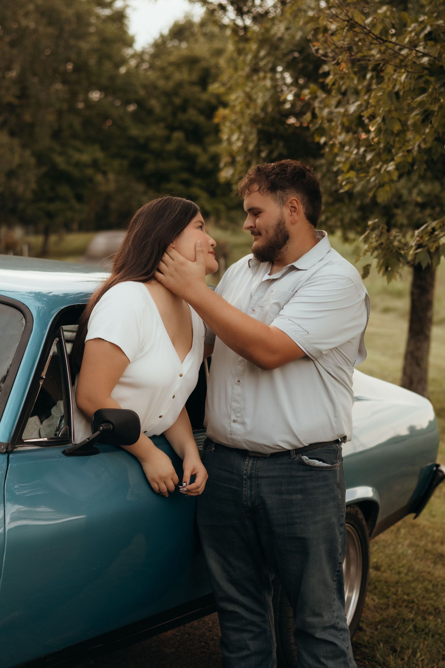 Couple sharing a romantic moment as a bearded man gently cradles a woman's chin beside a vintage blue car in a leafy outdoor setting.