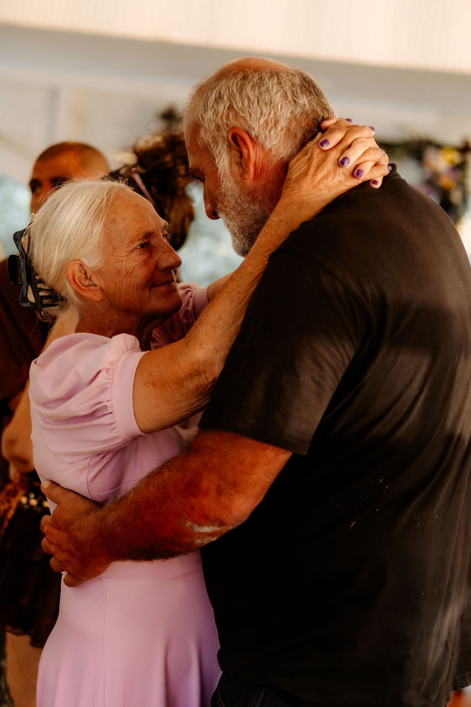Elderly couple sharing a tender embrace while slow dancing at a wedding celebration, the woman smiling in a lilac dress.