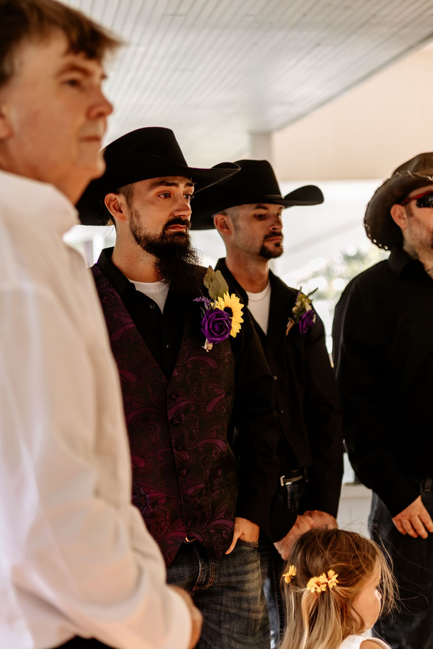Groomsmen in black cowboy hats and purple floral boutonnieres stand together at a wedding as a little girl looks on.