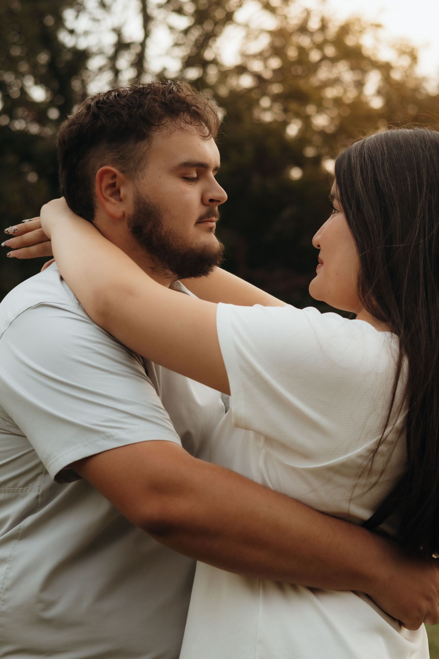 Couple embracing outdoors at golden hour, a bearded man in a gray shirt and a long-haired woman in white gazing at each other.
