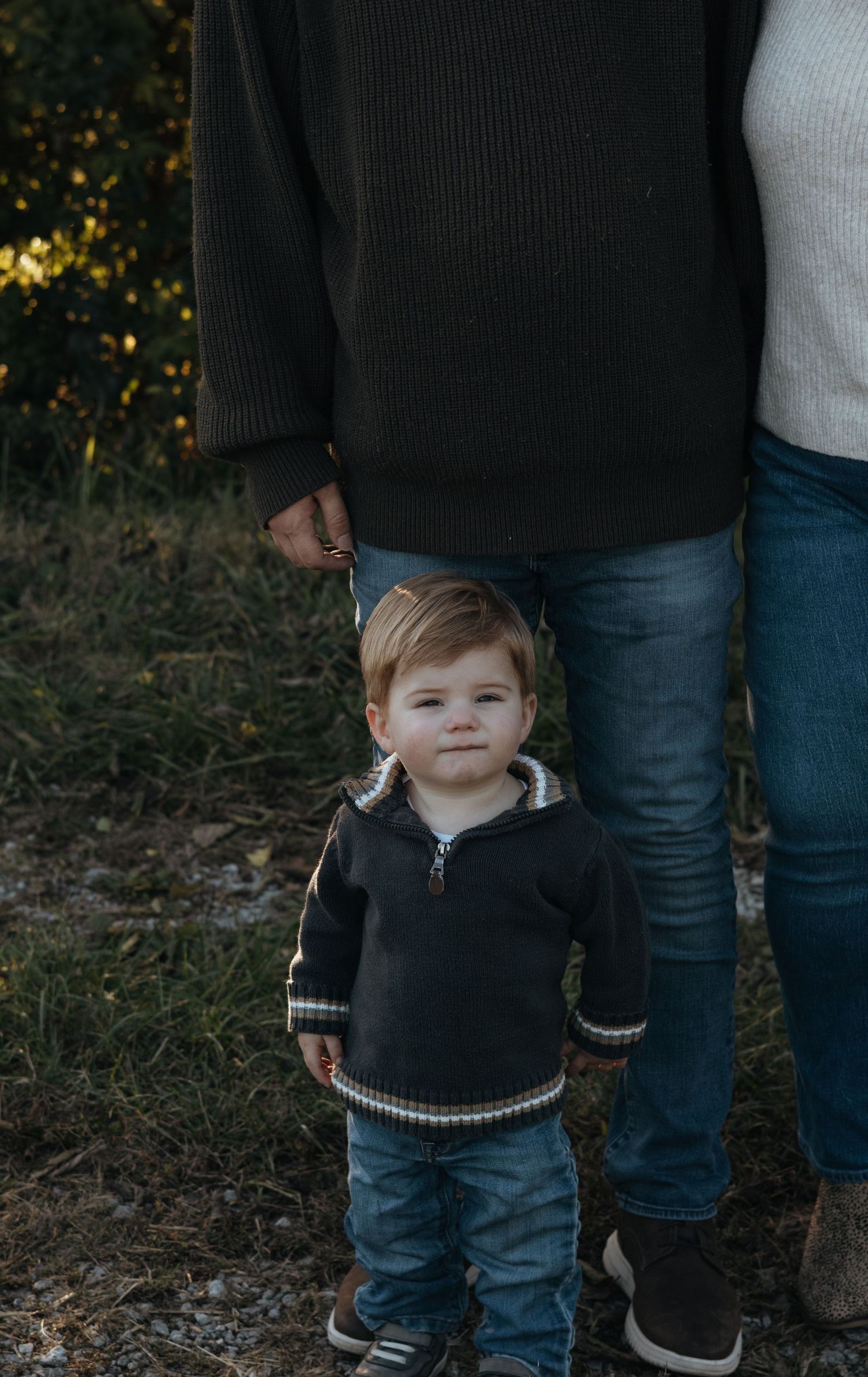 Toddler in a gray zip-up sweater and jeans stands on grass in front of two adults, with only their legs and torsos visible.