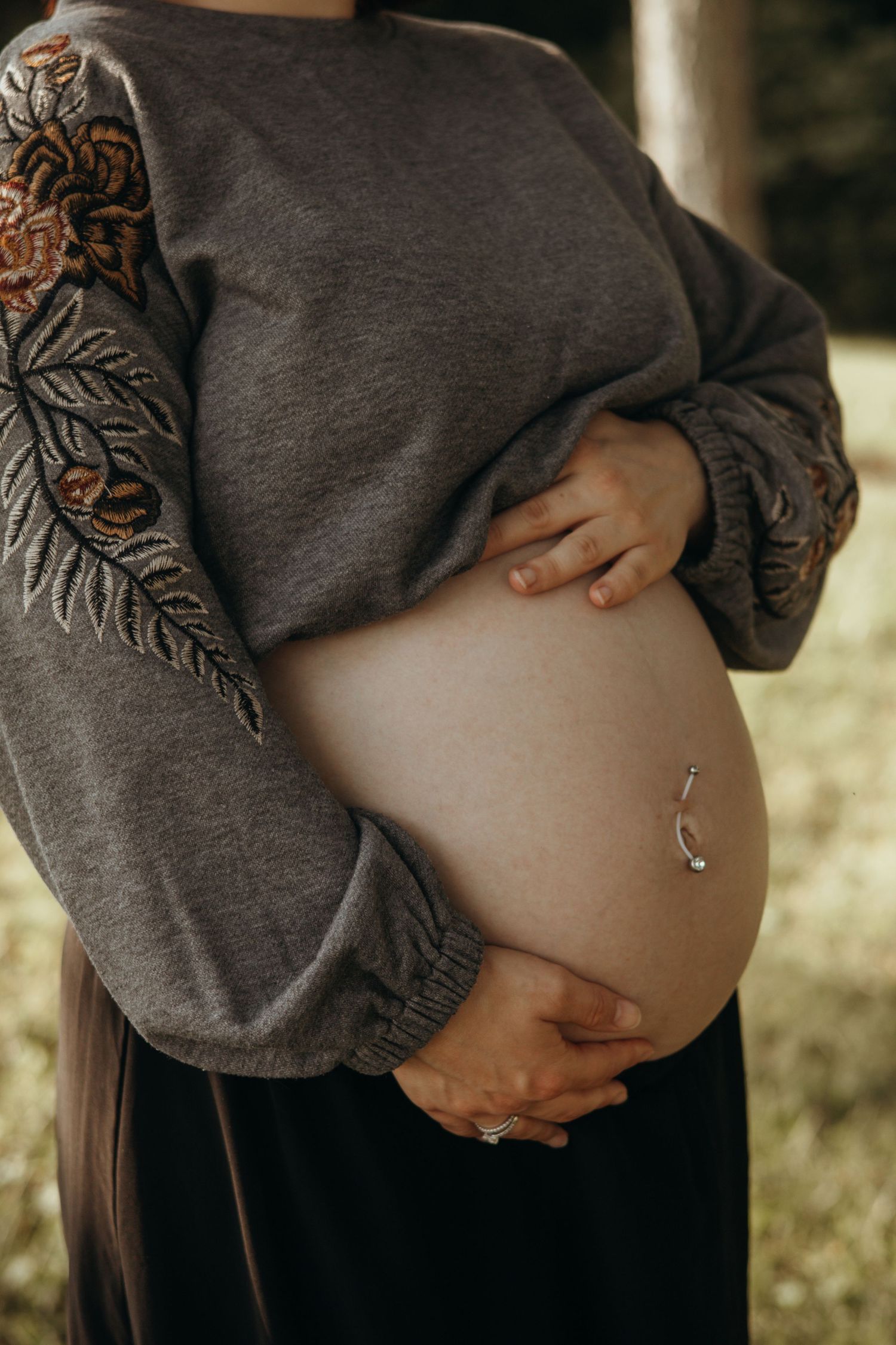 Pregnant woman in a gray embroidered sweater cradling her bare baby bump with a navel piercing outdoors in soft natural light.