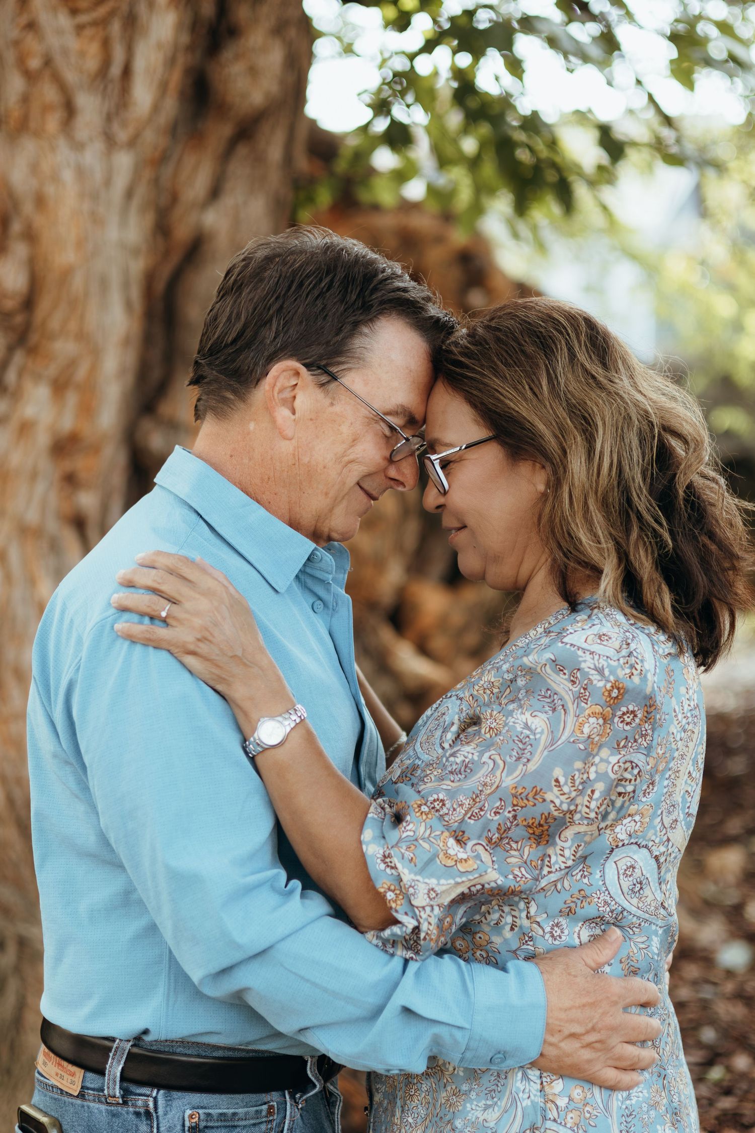 Middle-aged couple in glasses embracing forehead to forehead beside a large tree trunk, he in a blue shirt and she in a paisley blouse.