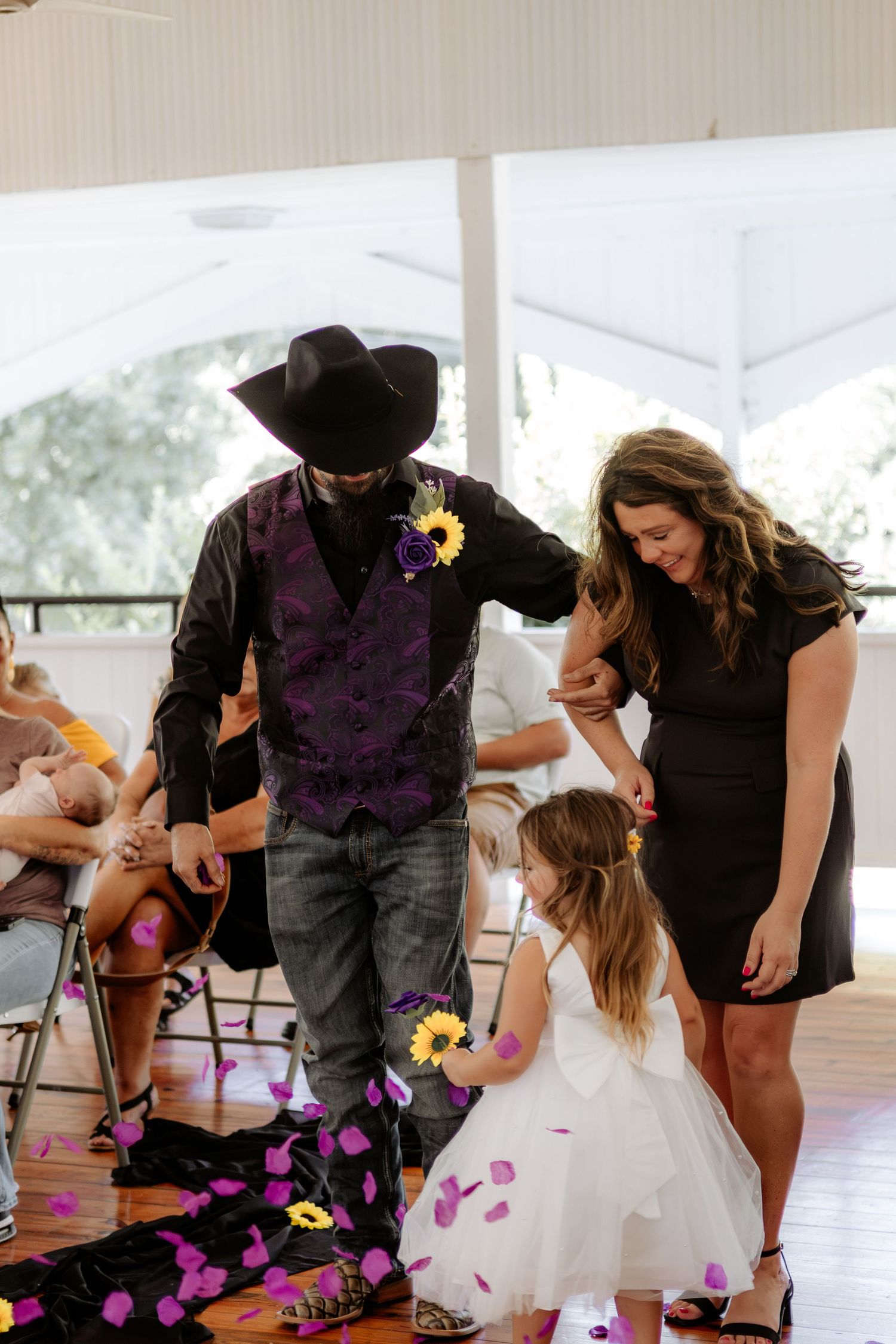 Groom in a black cowboy hat and purple vest walks down the aisle with a woman and flower girl as purple petals fall at a rustic wedding.