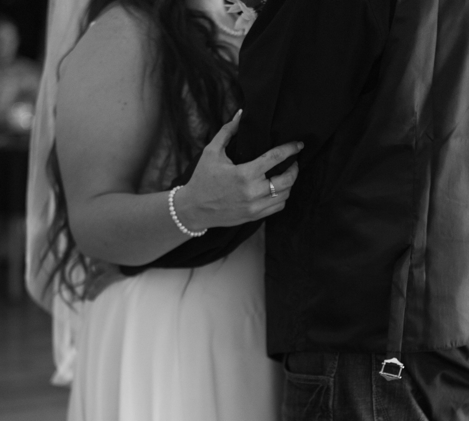 Black and white photo of a bride in a white dress and pearl bracelet embracing her partner in a dark jacket and jeans.